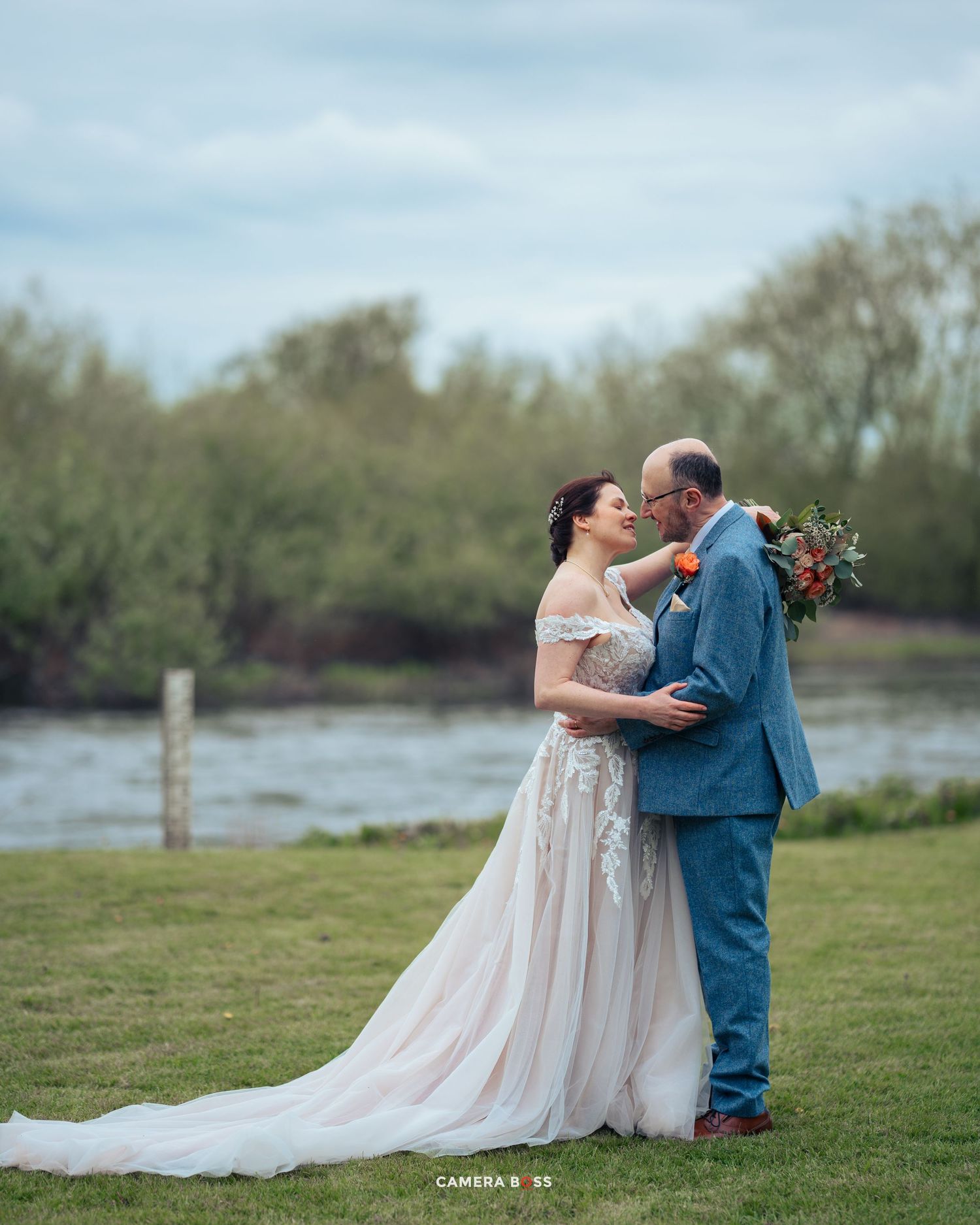 A couple embraces in a romantic lakeside wedding photo with the bride in a flowing off-shoulder gown and groom in blue suit.
