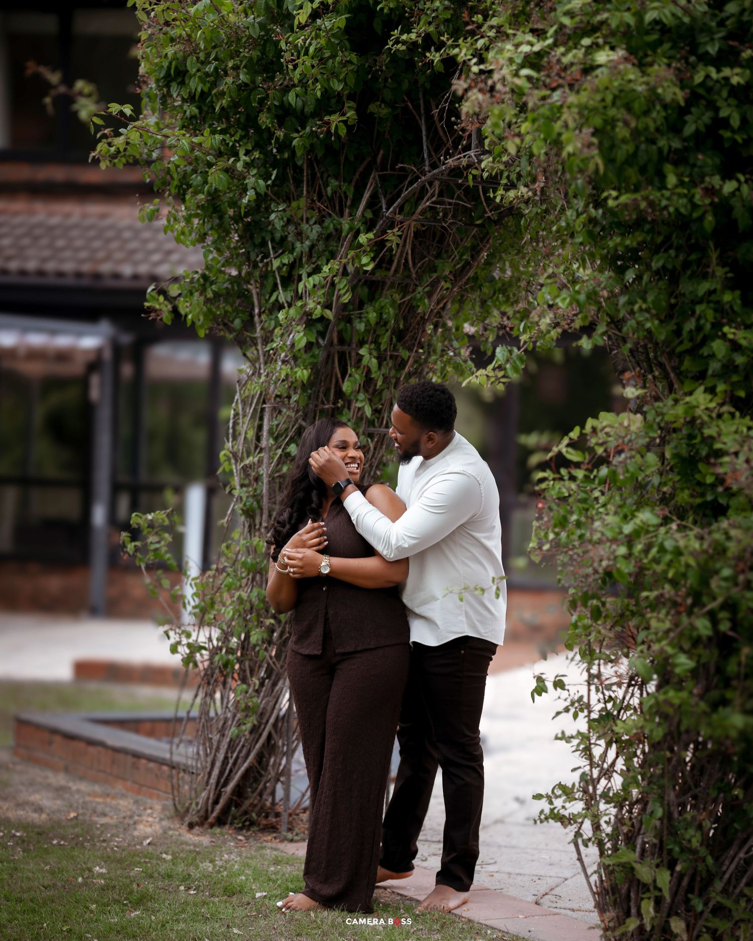 A romantic couple embraces on a garden pathway surrounded by flowering vines and greenery.