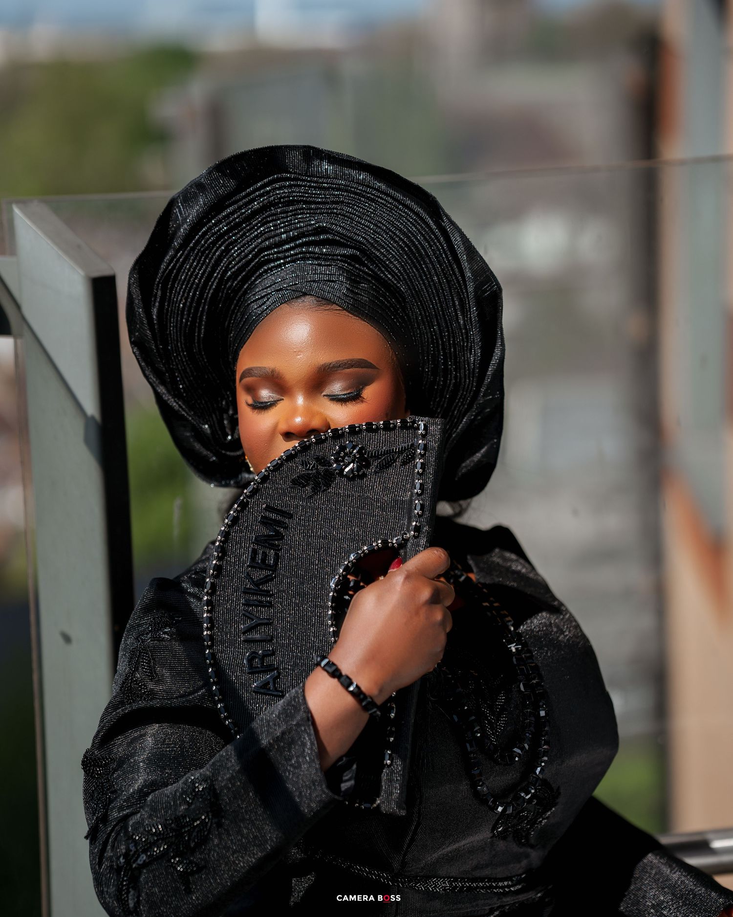 Person wearing elegant black traditional Nigerian attire with gele headwrap poses with decorative black handheld fan.