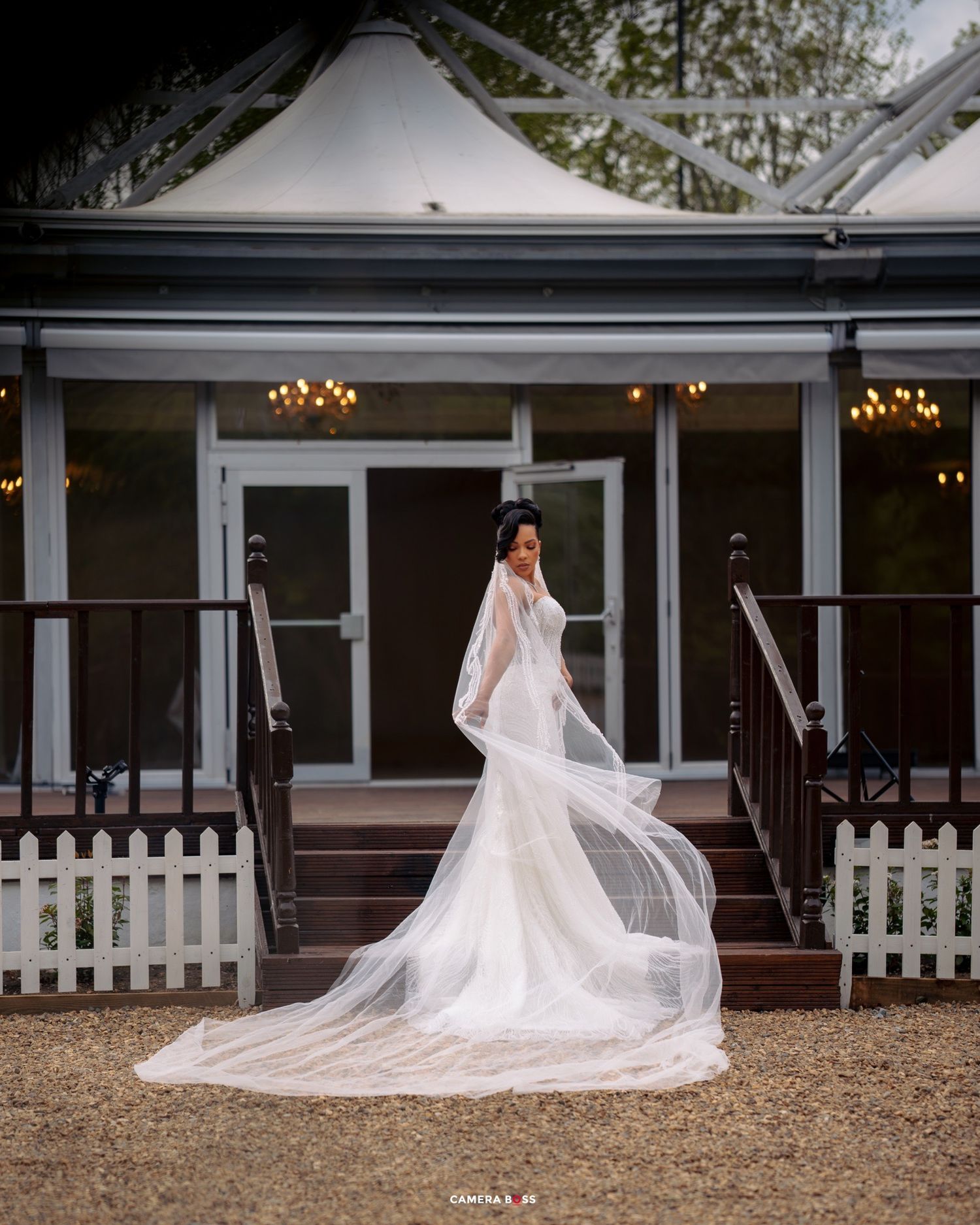 Elegant bride in form-fitting lace gown with flowing cathedral veil poses on porch with white picket fence and chandeliers.