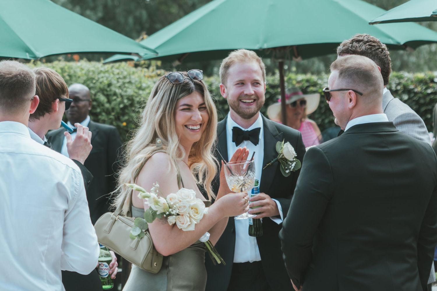 Wedding guests socializing and enjoying drinks under green patio umbrellas at an outdoor reception.