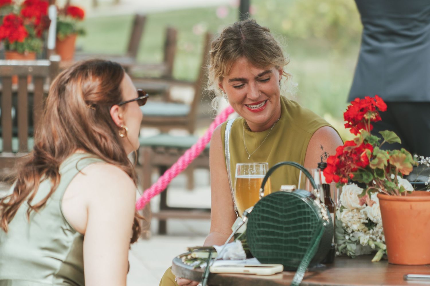 Two guests share laughter over drinks at an outdoor table decorated with red flowers.