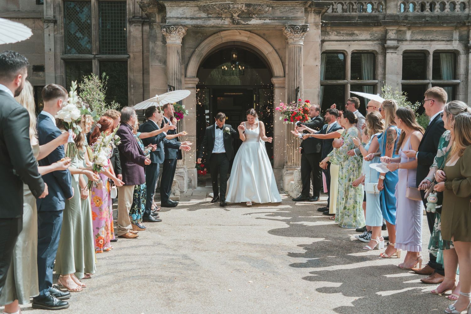 Wedding guests line the entrance of a historic stone building throwing confetti as newlyweds exit the ceremony.
