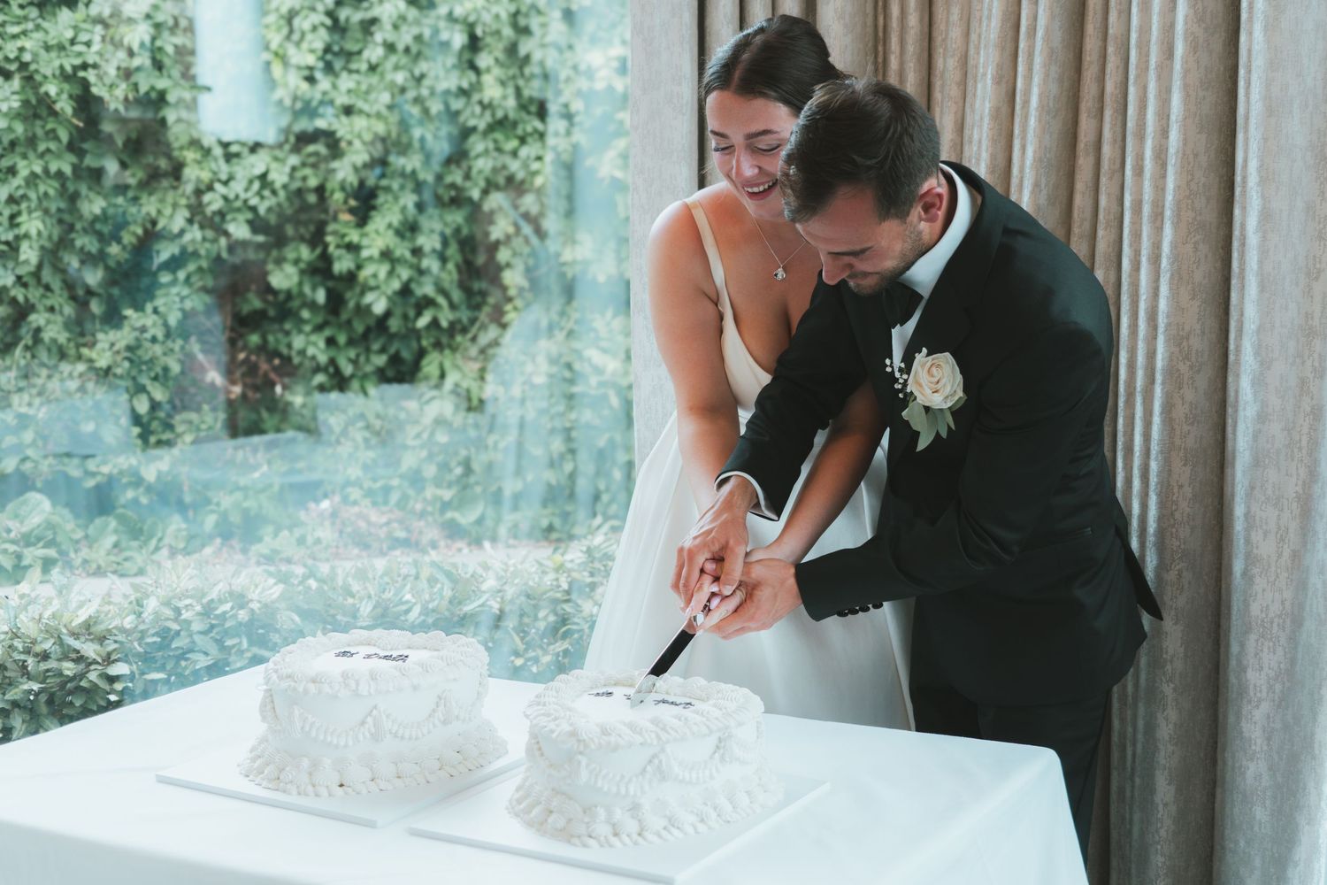 Newlyweds cutting their white wedding cake together by window.