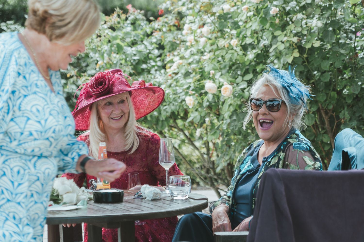 Wedding guests enjoy drinks and conversation at an outdoor garden table with colorful sun hats.