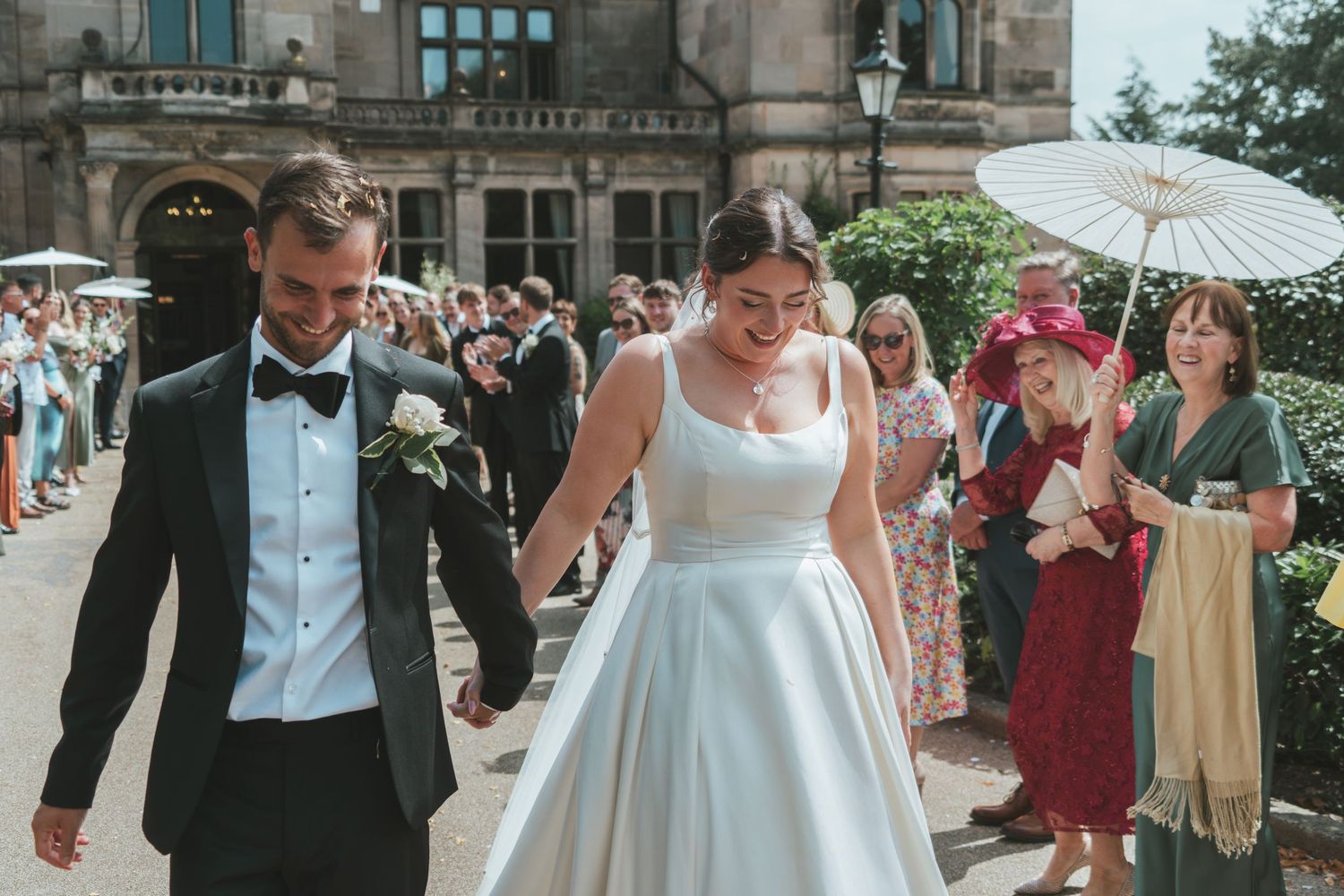Wedding party enjoying candid moments outside stately building with parasol.