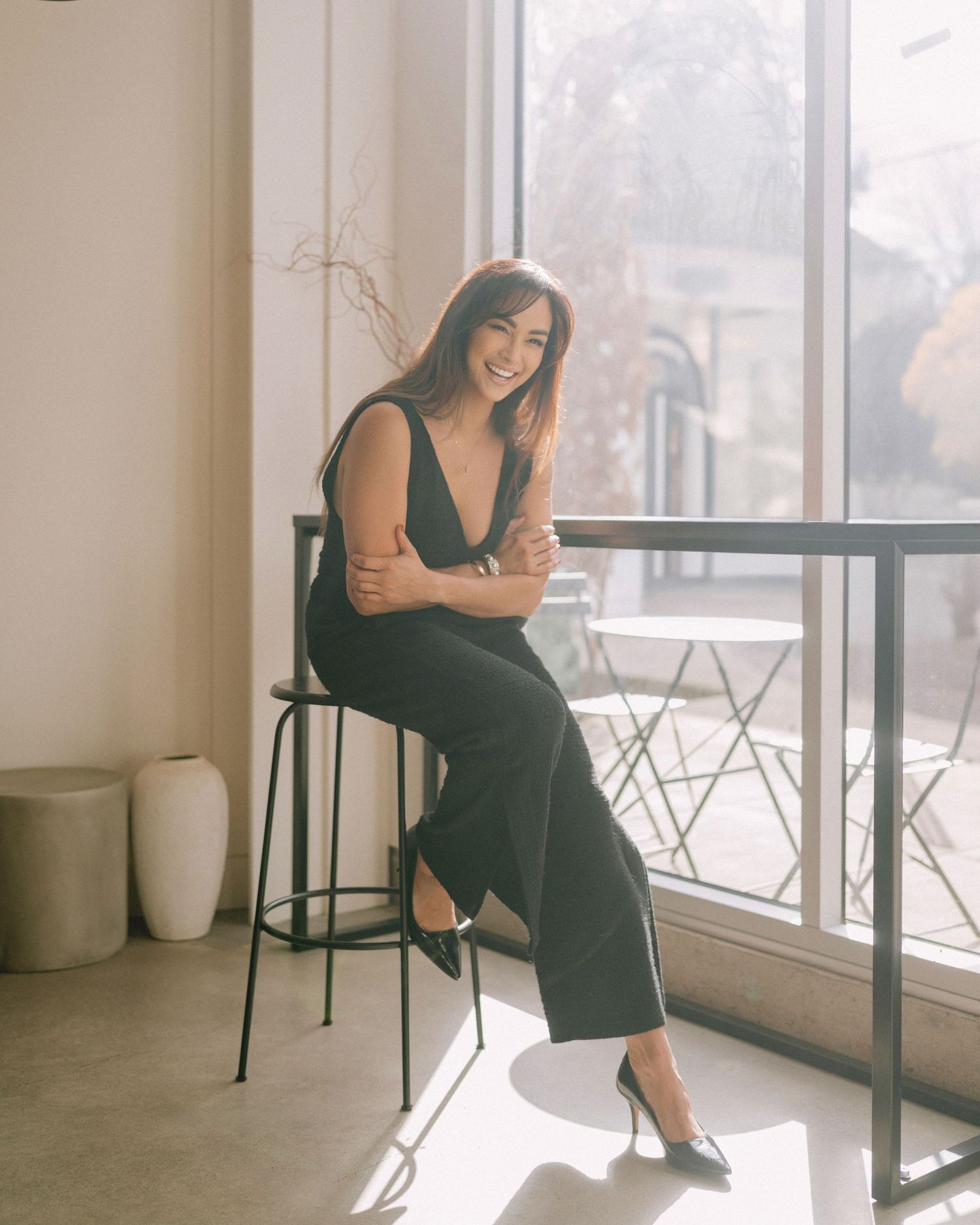 Woman in black outfit sits at high table by window in minimalist interior.