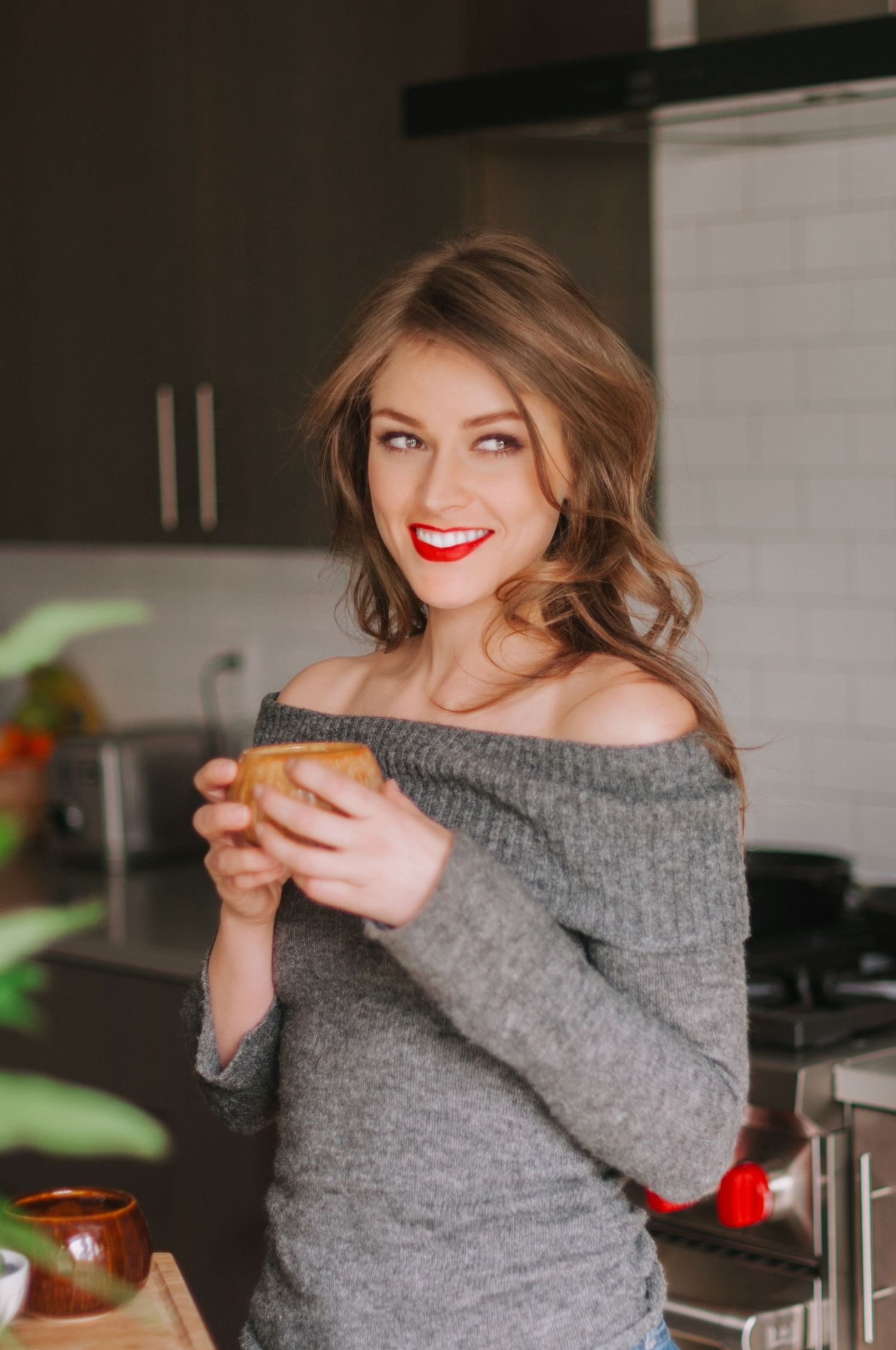 Person in off-shoulder gray sweater holds mug in kitchen with joyful expression.