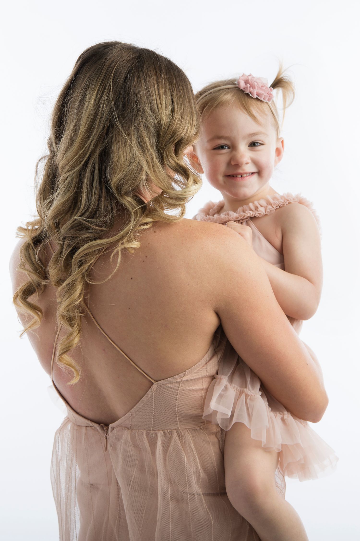 A heartwarming moment in blush pink dresses as a woman with wavy blonde hair holds a smiling small child against white backdrop.