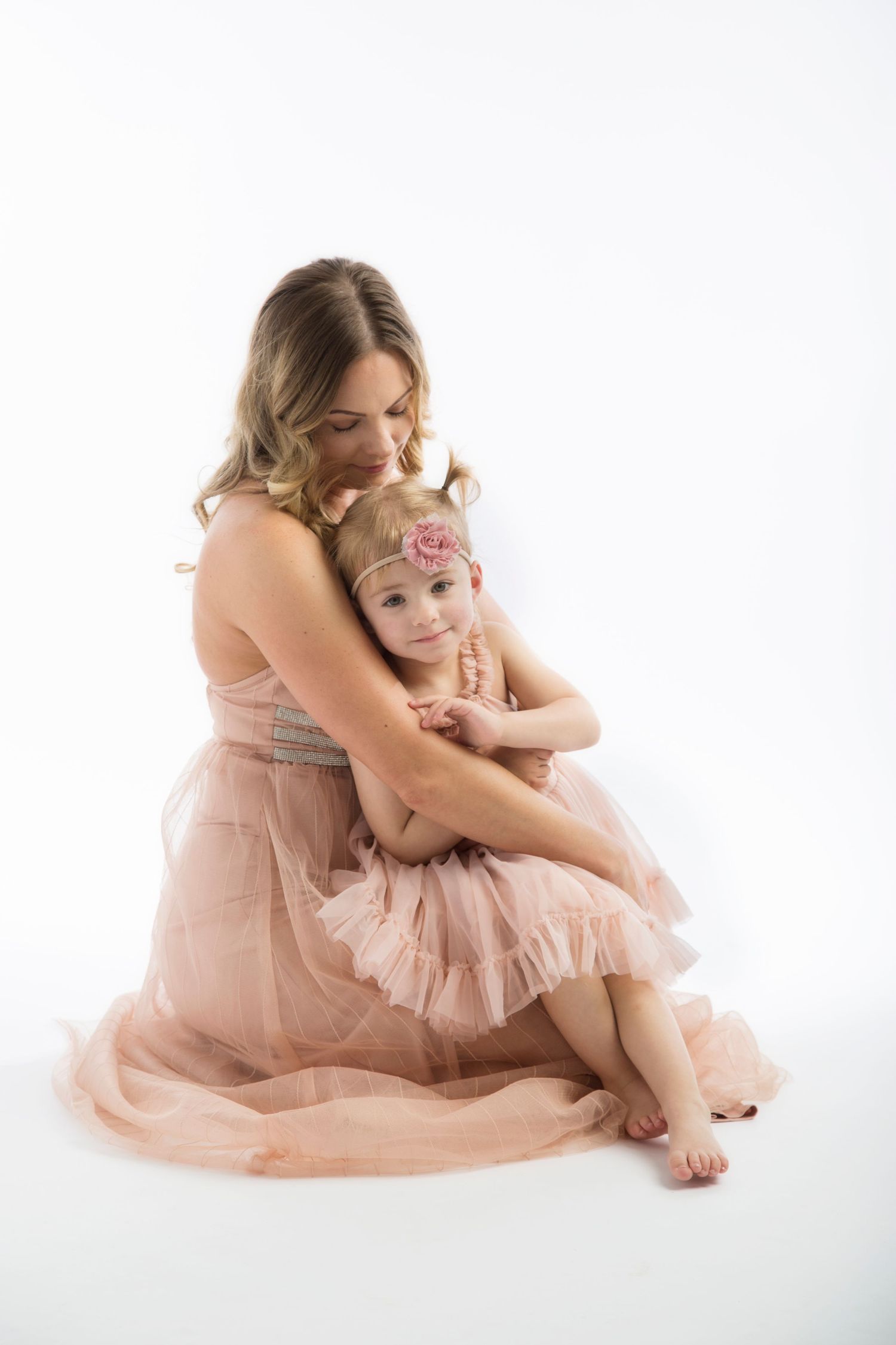 A mother and daughter in pink dresses embrace tenderly while sitting on a white backdrop in a studio setting.