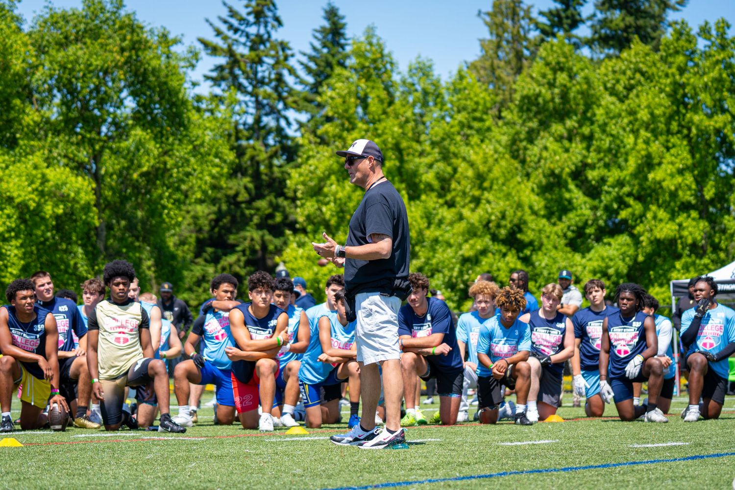 A coach instructs athletes during an outdoor sports training session on a sunny day with green trees in background.