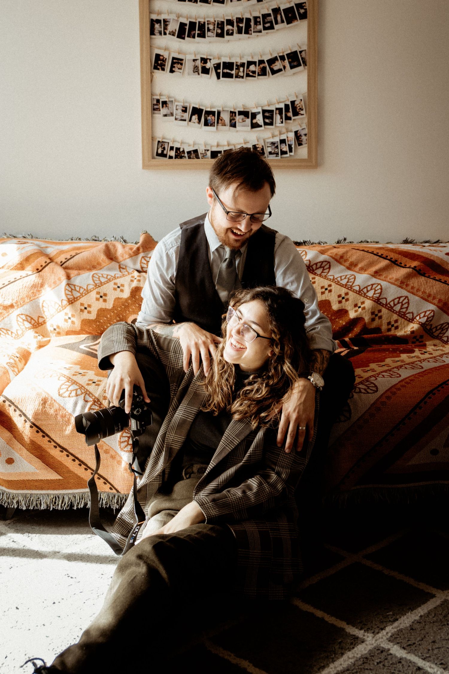 Vintage-styled couple lounging on patterned couch beneath wall of polaroid photos.