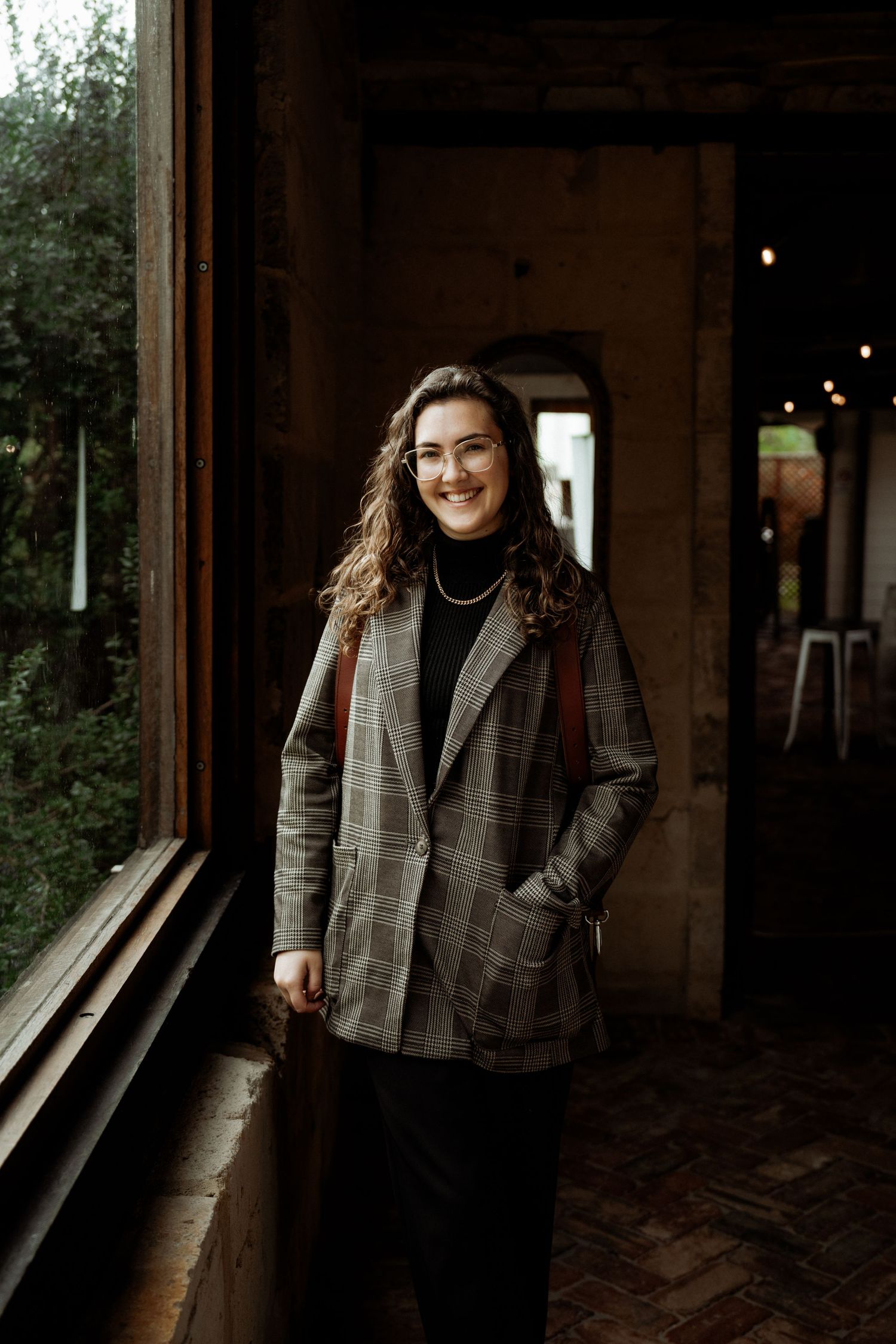 Wearing a plaid blazer and black turtleneck, someone poses by a rustic window with natural lighting indoors.