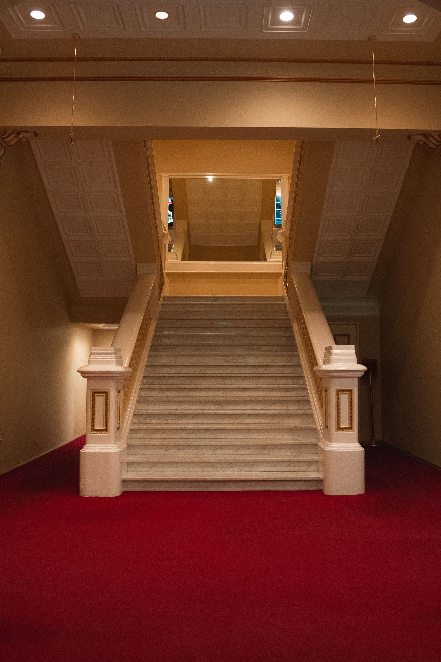 Elegant marble staircase with red carpet and cream walls leads upward in a grand interior space with recessed lighting.