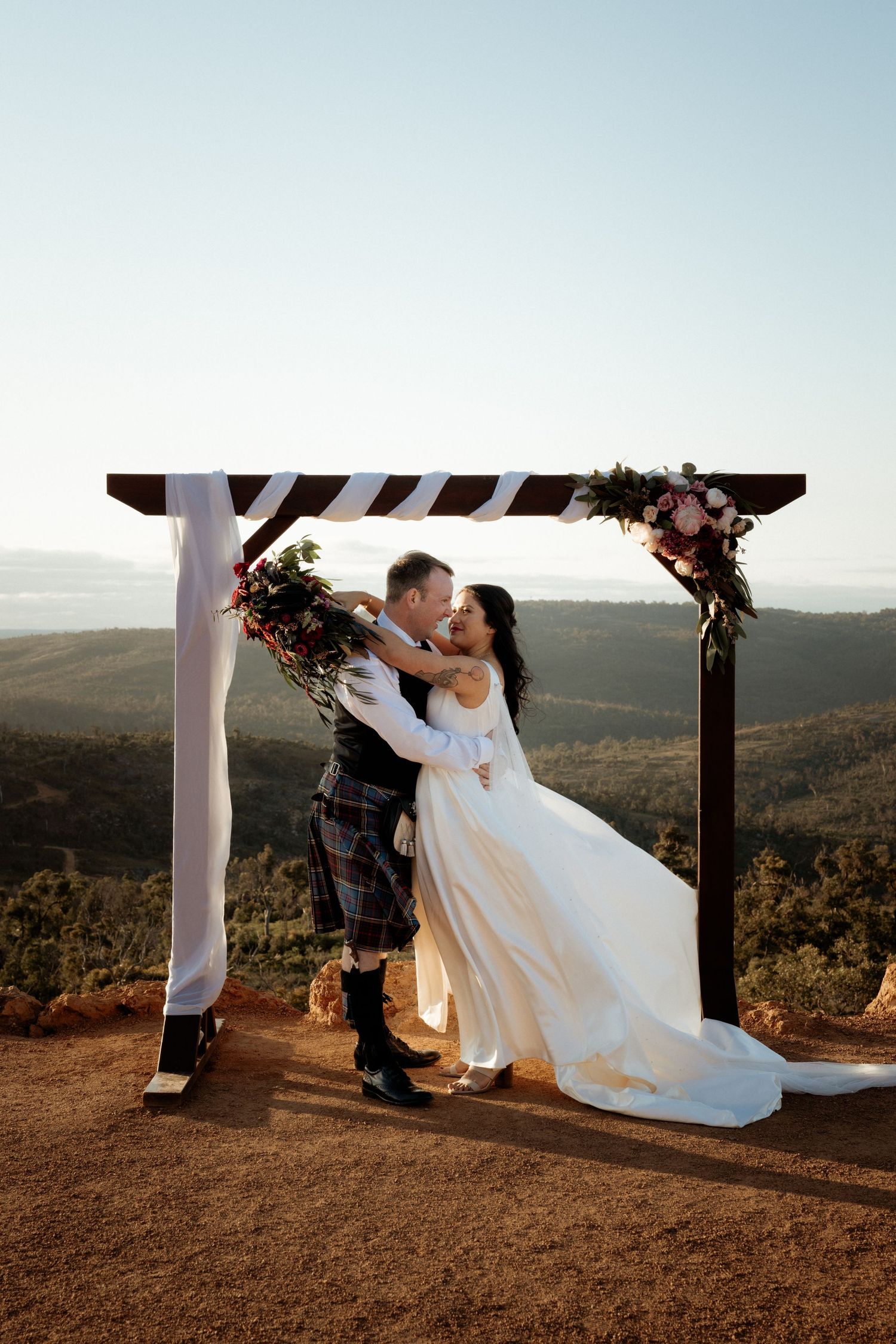 Wedding couple embracing under floral arch with mountain vista backdrop at sunset.