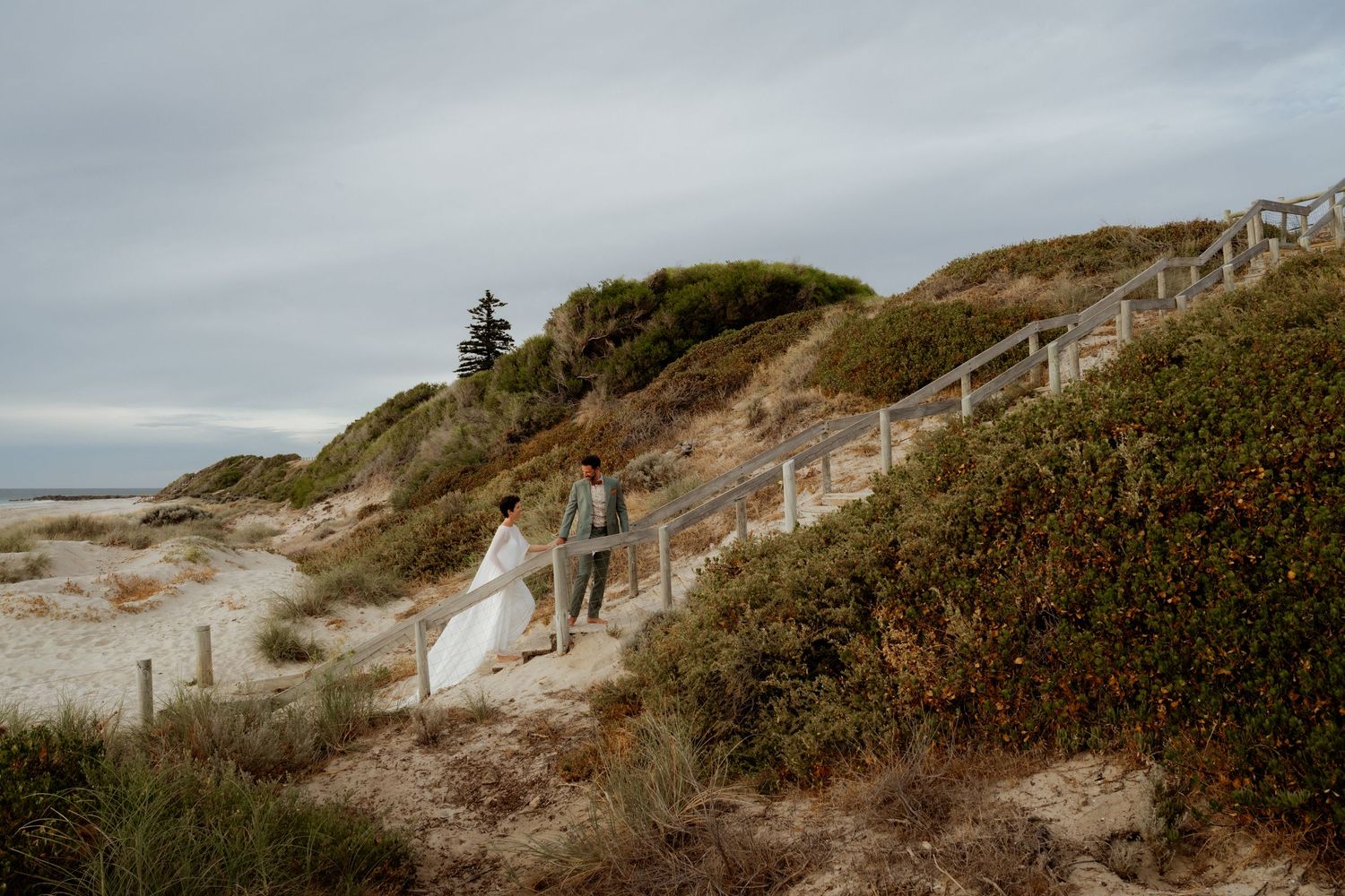 A couple walking down wooden stairs on a scenic coastal dune path.