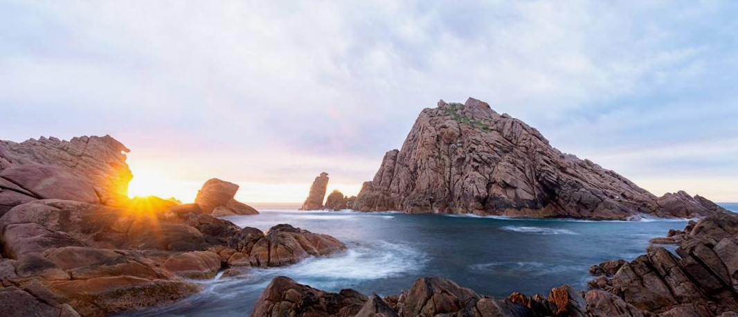 Dramatic panorama of rocky coastline with sunburst through granite formations at sunrise.