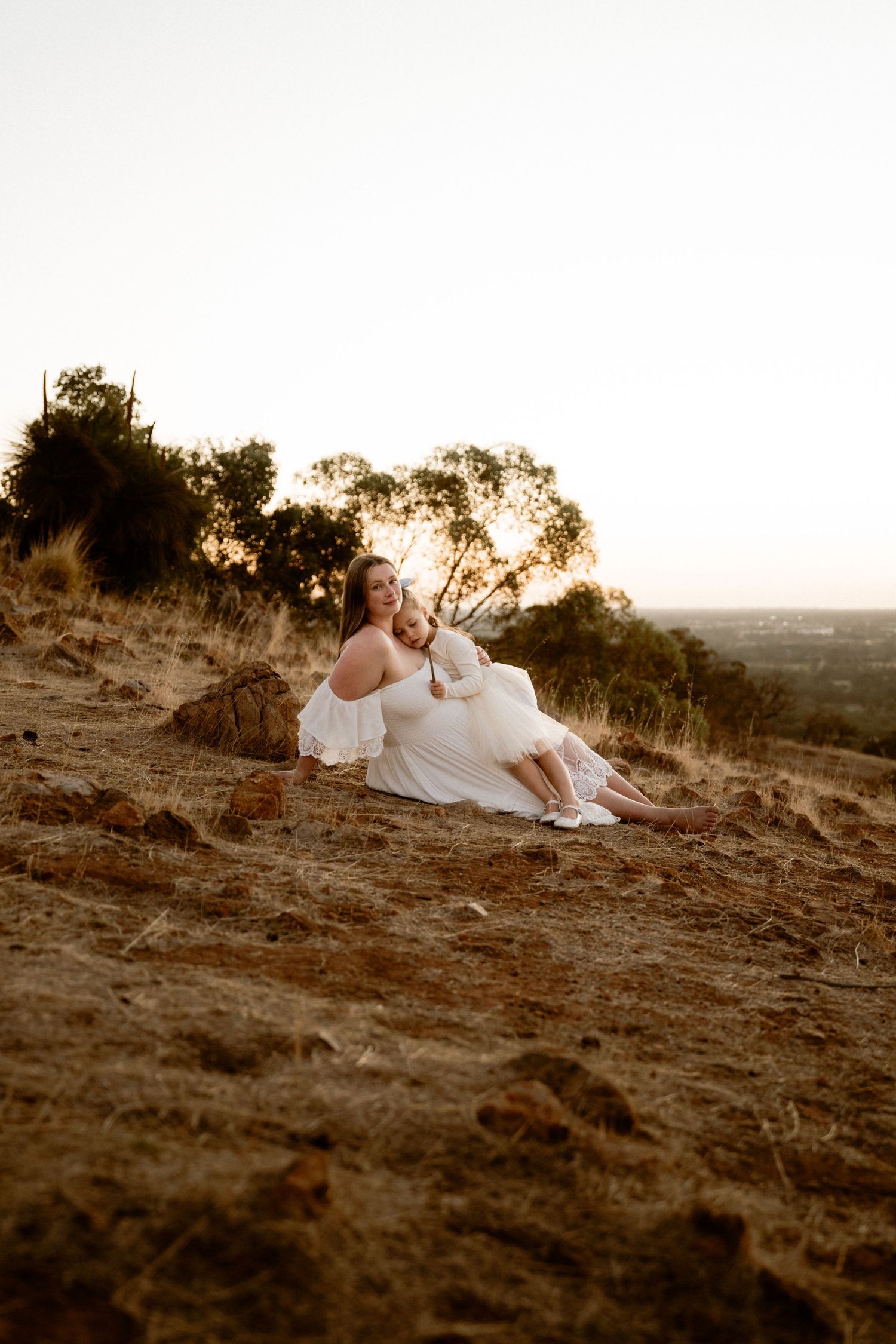 A woman in white dress sits on hillside during golden hour sunset.