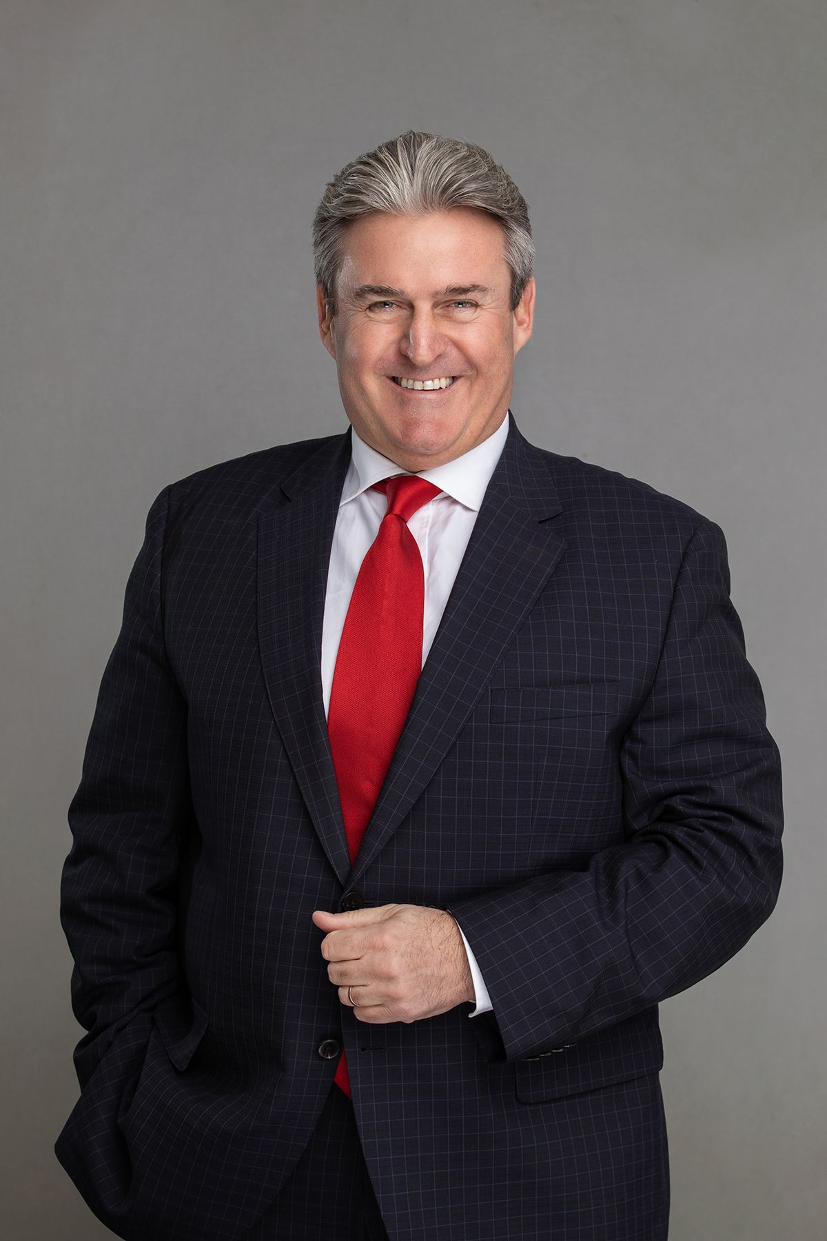Professional studio portraits of someone wearing a dark suit and red tie against a gray background.