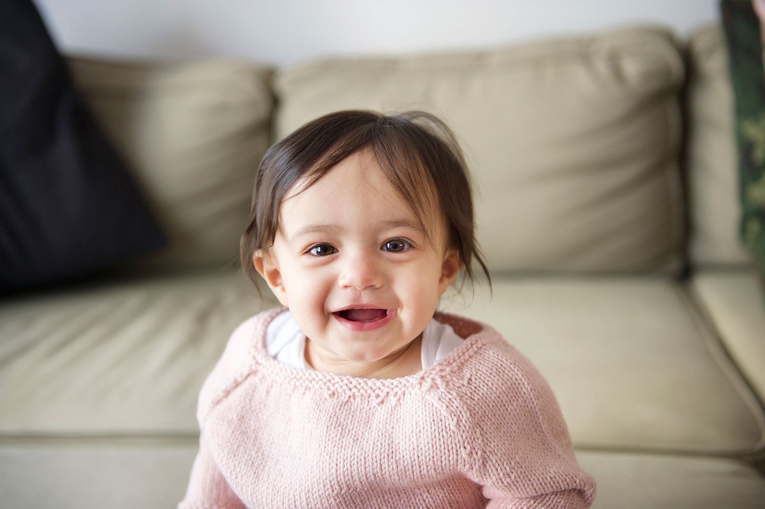 Baby wearing pink knit sweater sitting and smiling on beige couch. Captured by Toronto based baby and family photographer, Phoebe Lee Photo.