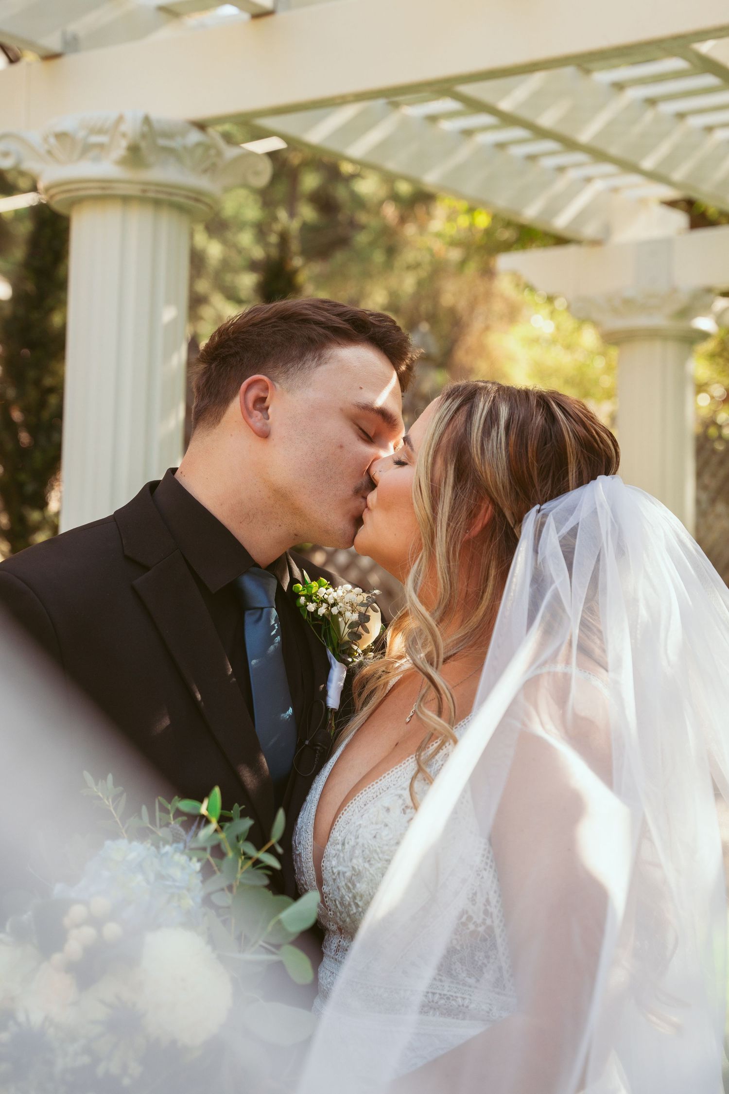 A romantic wedding moment captured under a white pergola with soft sunlight and floral details.