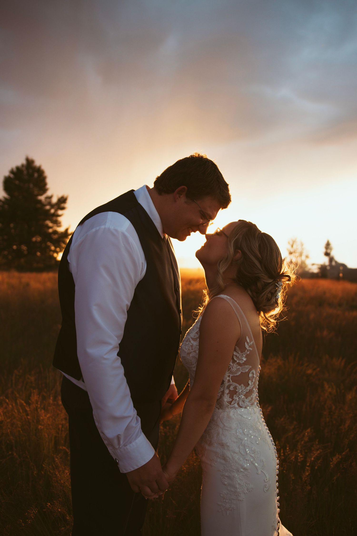 Romantic silhouette of a couple embraces during golden sunset in a meadow with lace wedding dress details.