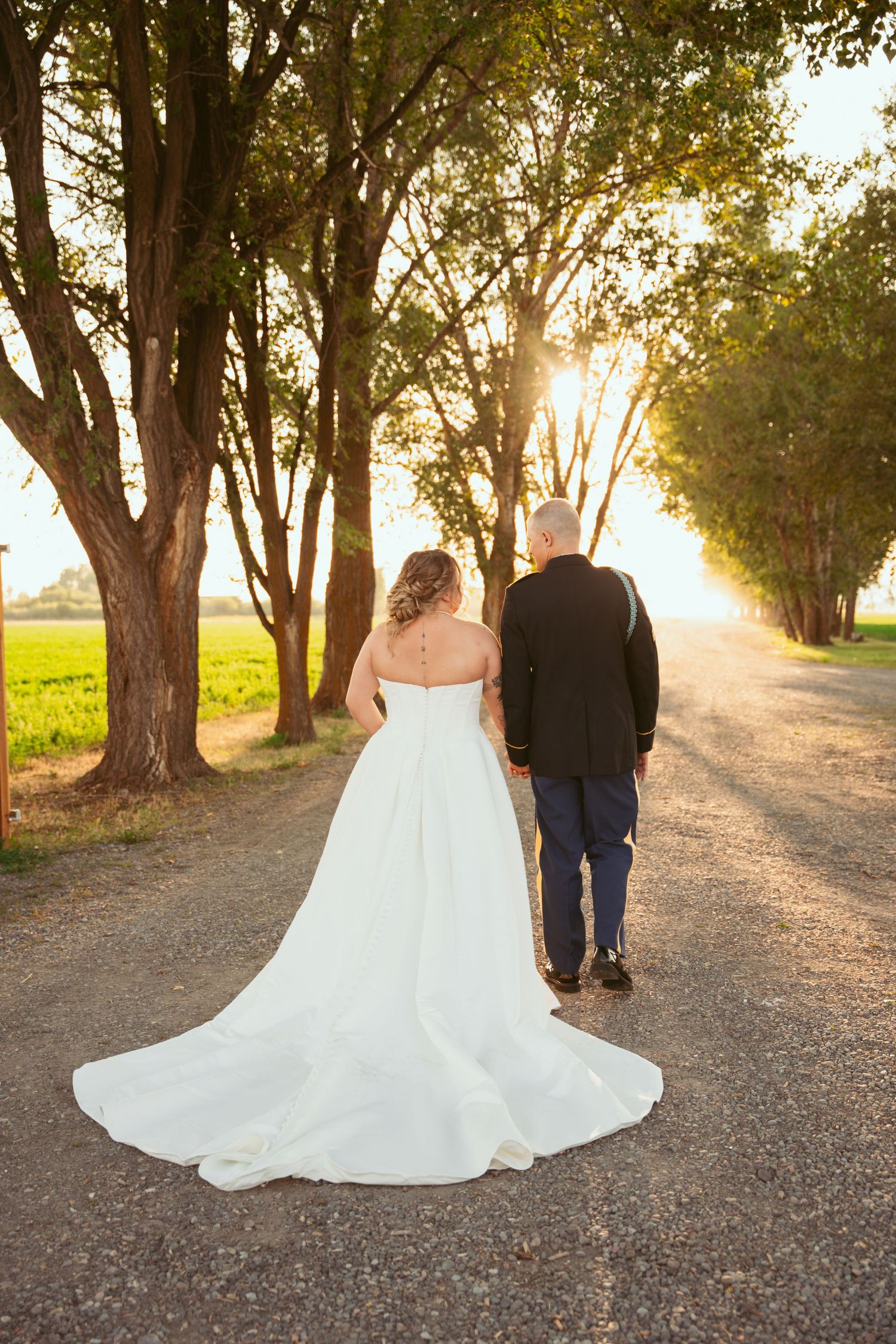 A couple walks hand in hand down a tree-lined dirt road at sunset, with the bride wearing a flowing white wedding gown.