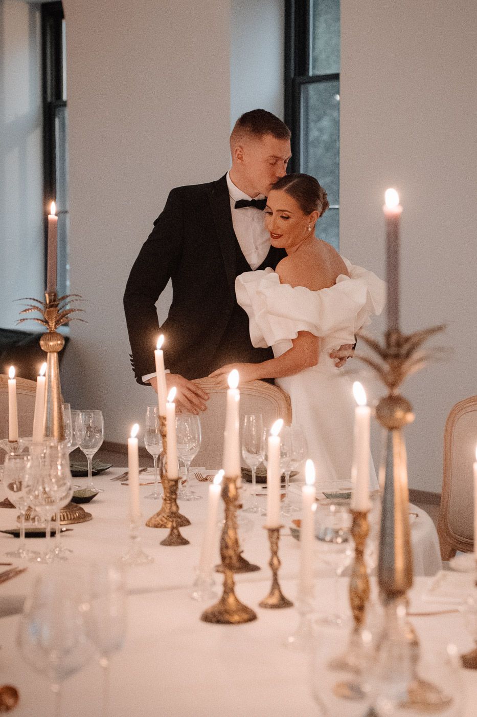 A romantic candlelit table setting with lit tapered candles in brass holders and wine glasses at a wedding reception.