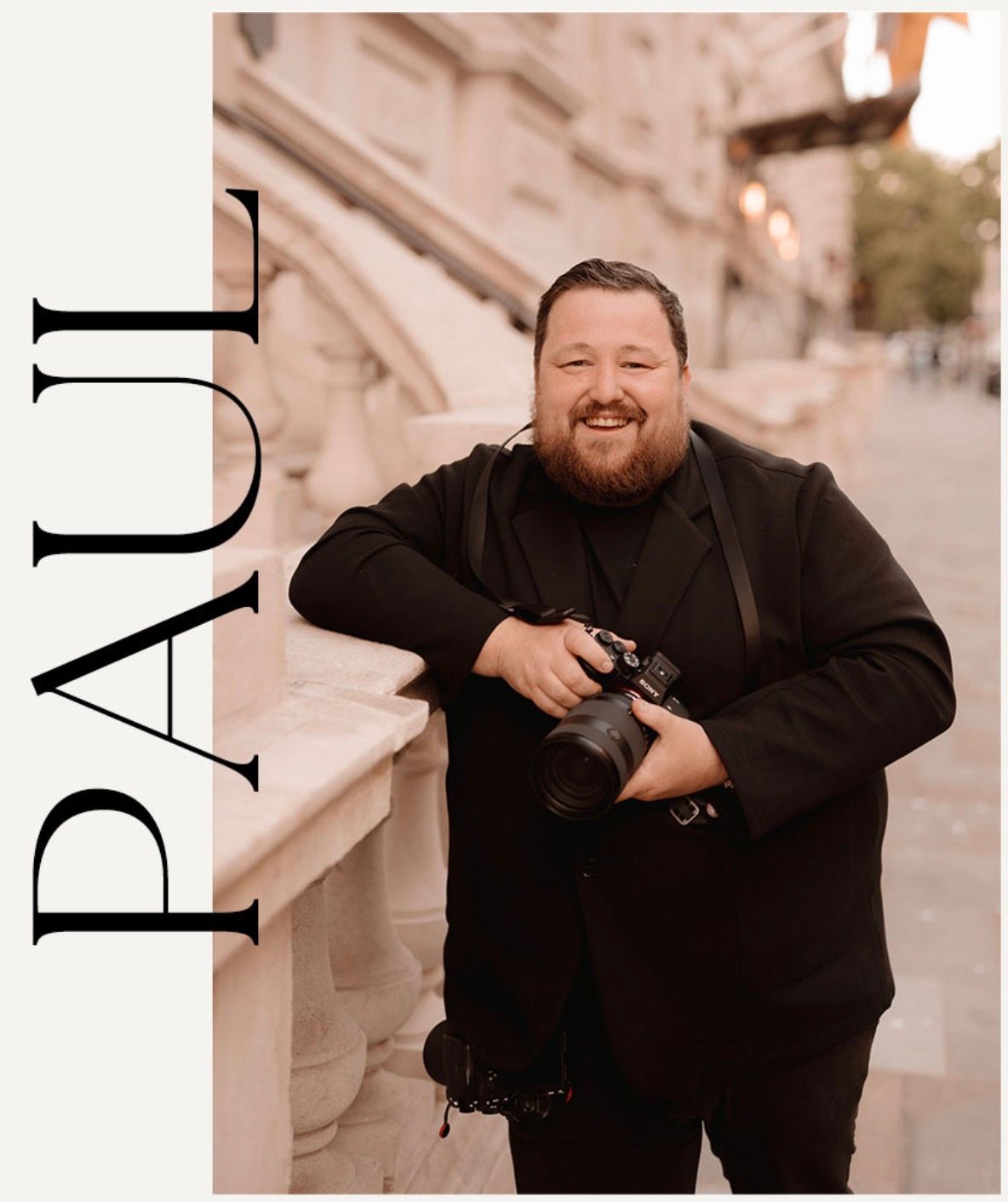 Professional photographer holding a digital camera while posing outdoors in a black outfit.