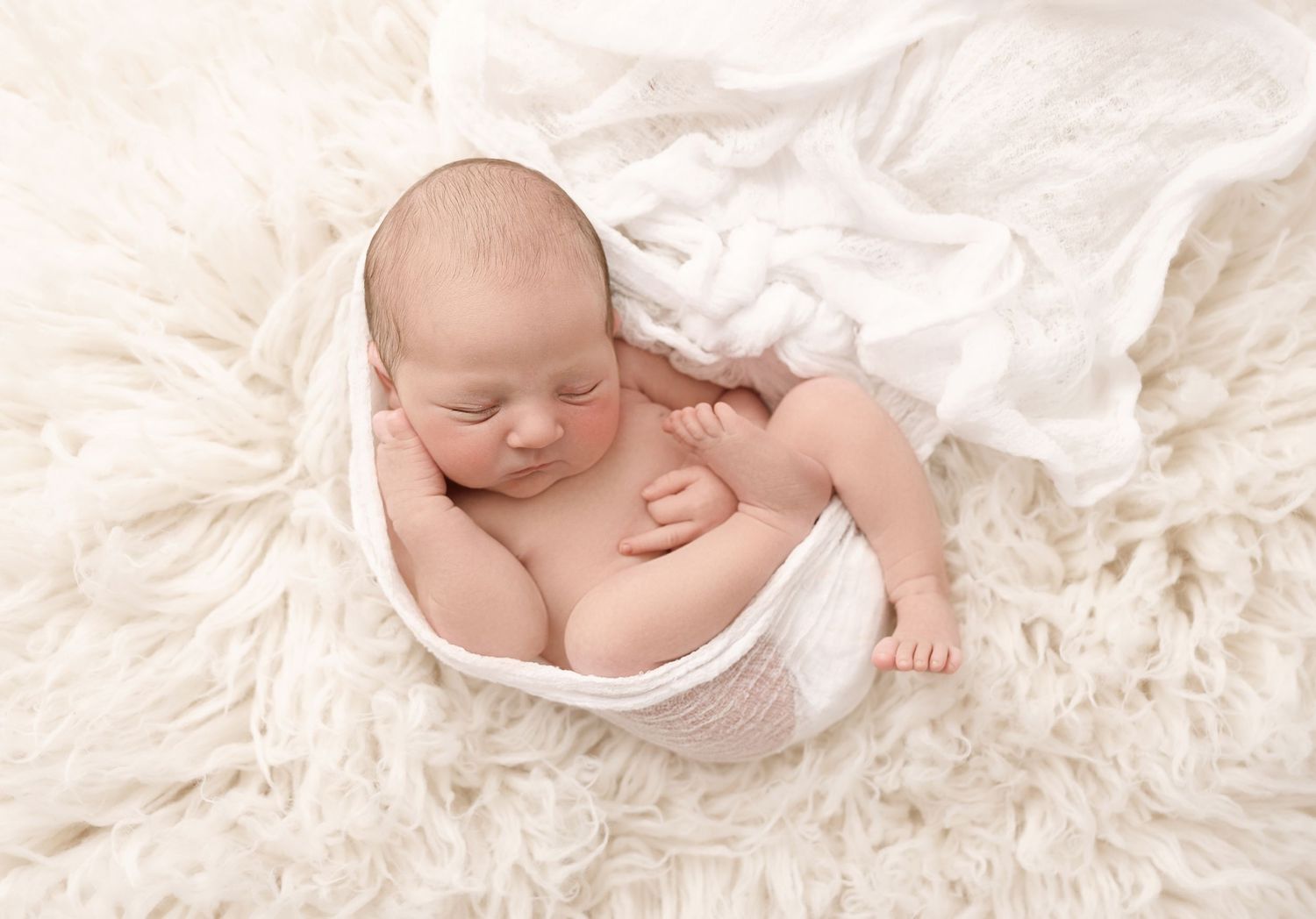 Peaceful sleeping newborn baby wrapped in white fabric nestles on soft cream textured blanket.