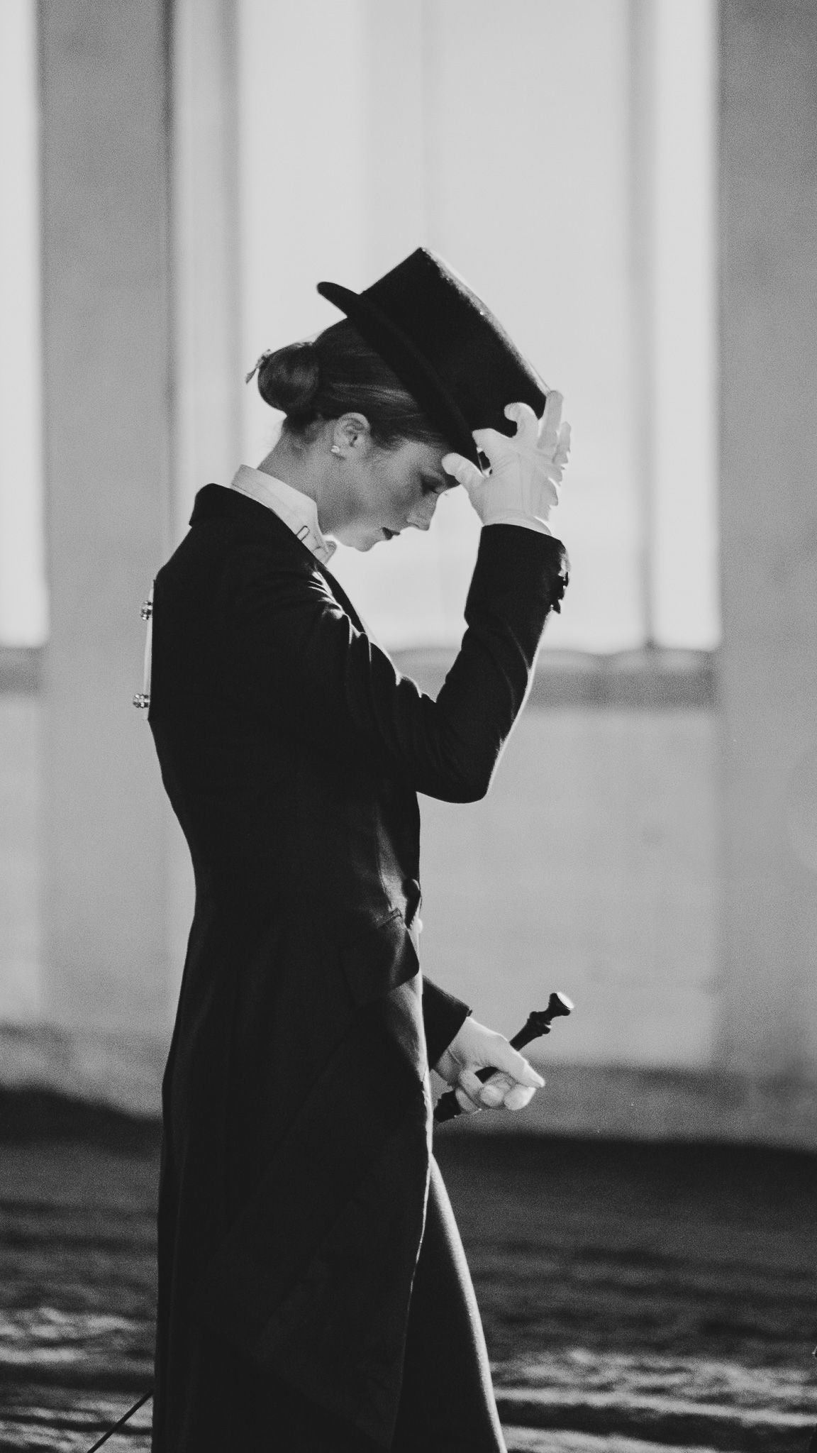 A figure in a vintage black suit and hat stands in profile against a light background in a black and white photograph.