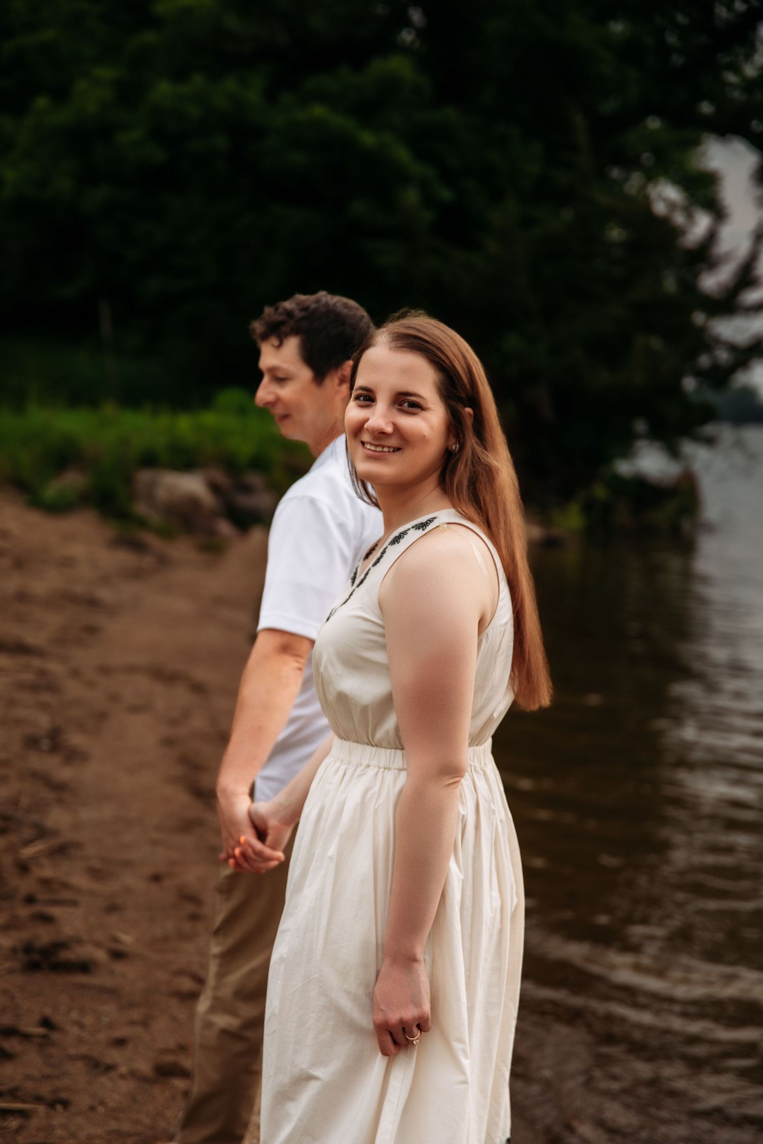 Couple in white outfits smiling and walking along the water's edge at sunset.