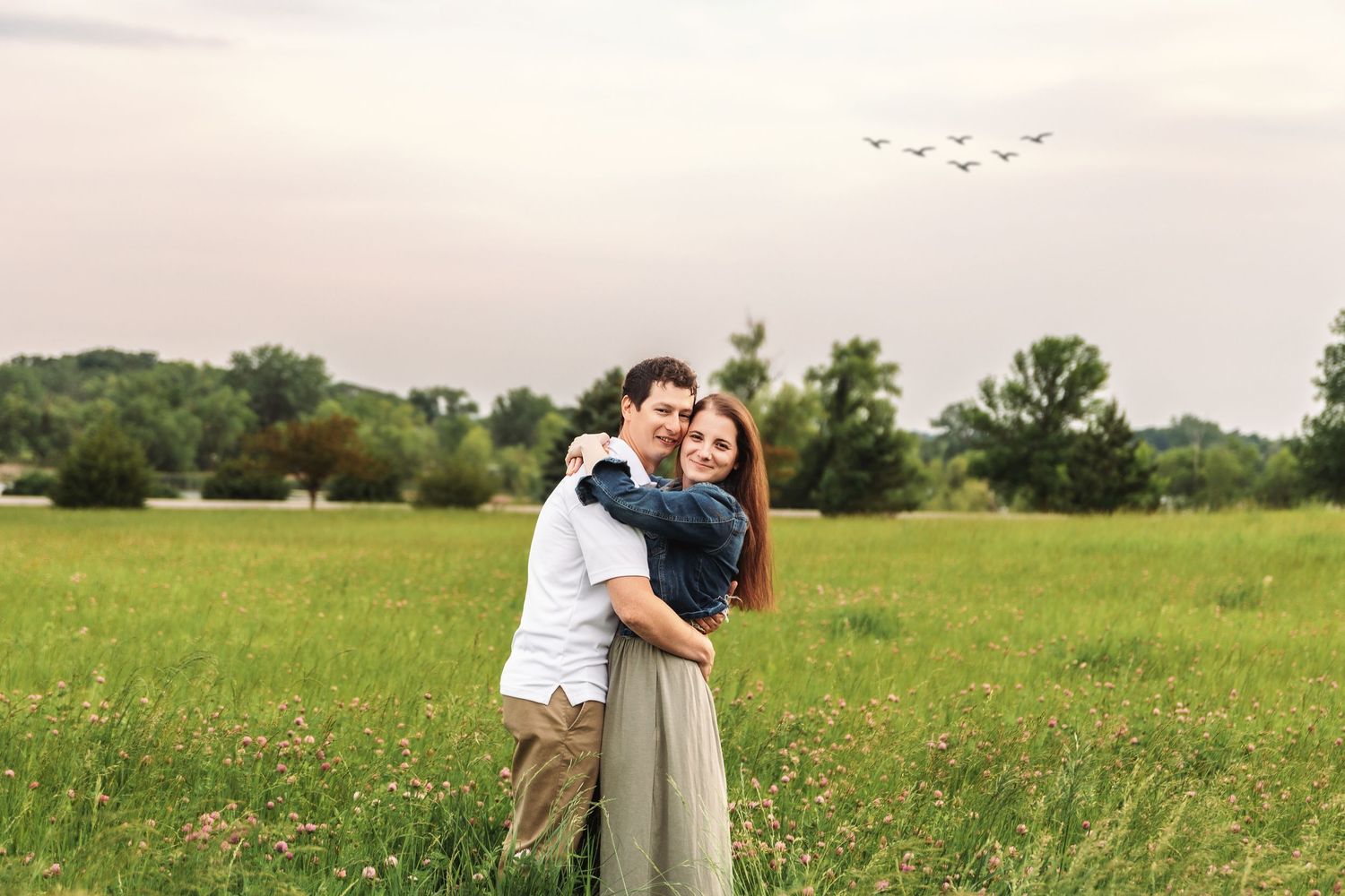 A couple embraces in a serene green meadow during golden hour as birds fly overhead in the sky.