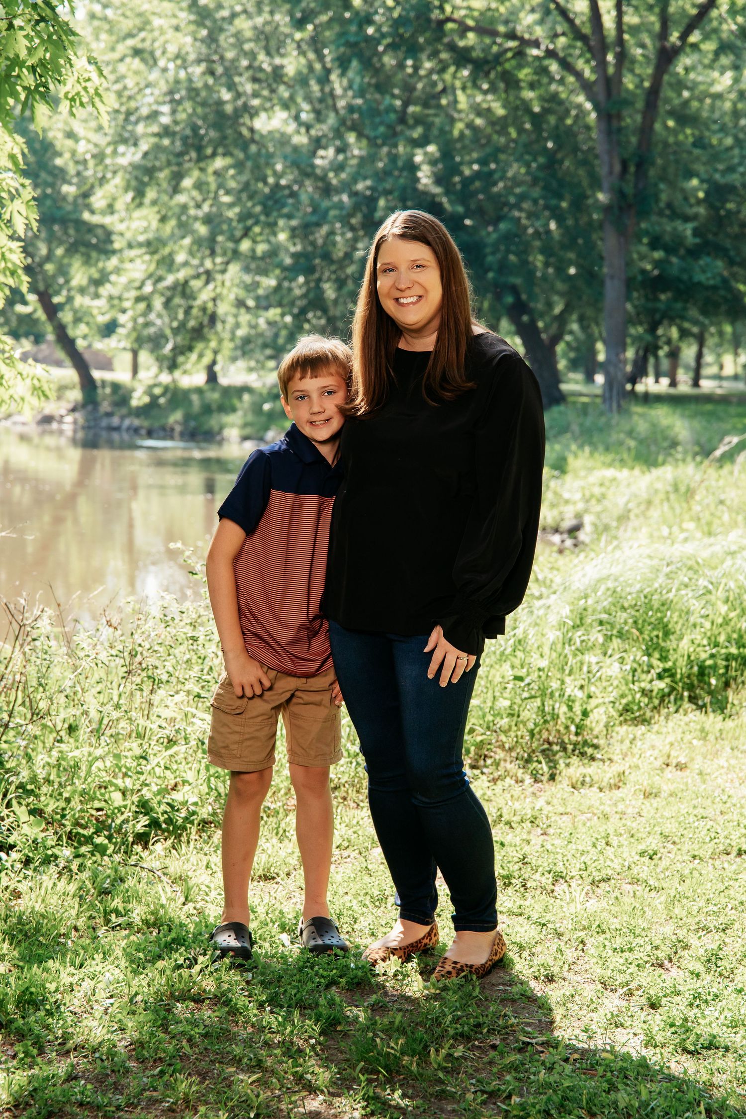 A parent and child share a joyful moment while standing together on grassy riverbank in a sunlit park.