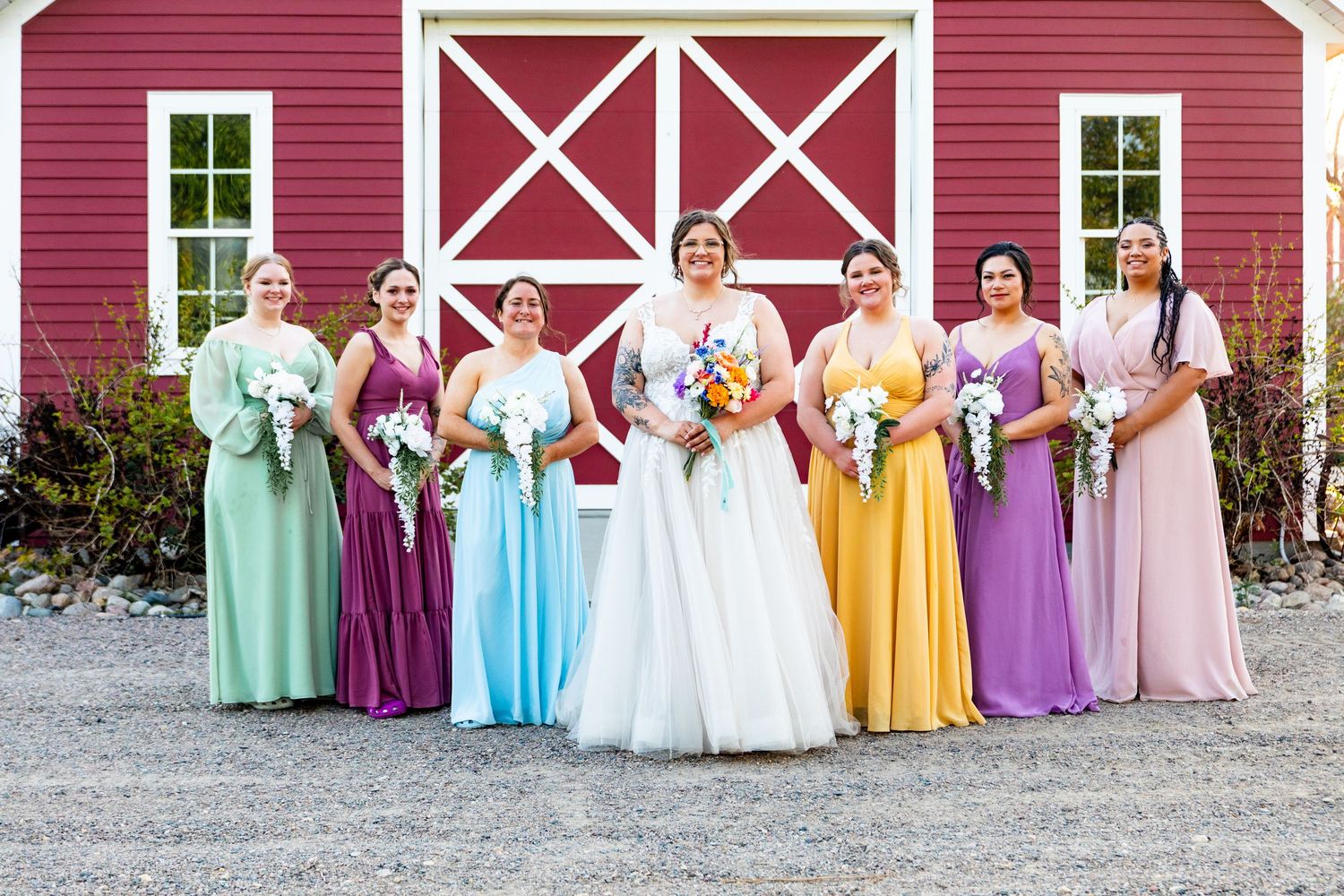 Wedding party in colorful dresses posed in front of a red barn for group photos.