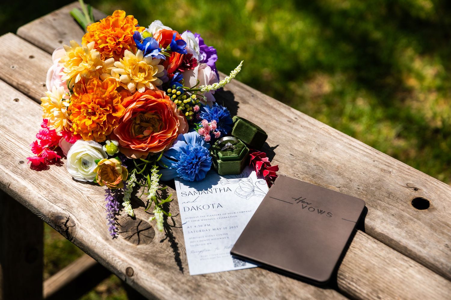Vibrant wedding bouquet with orange, blue and pink flowers sits next to a small book on wooden surface outdoors.