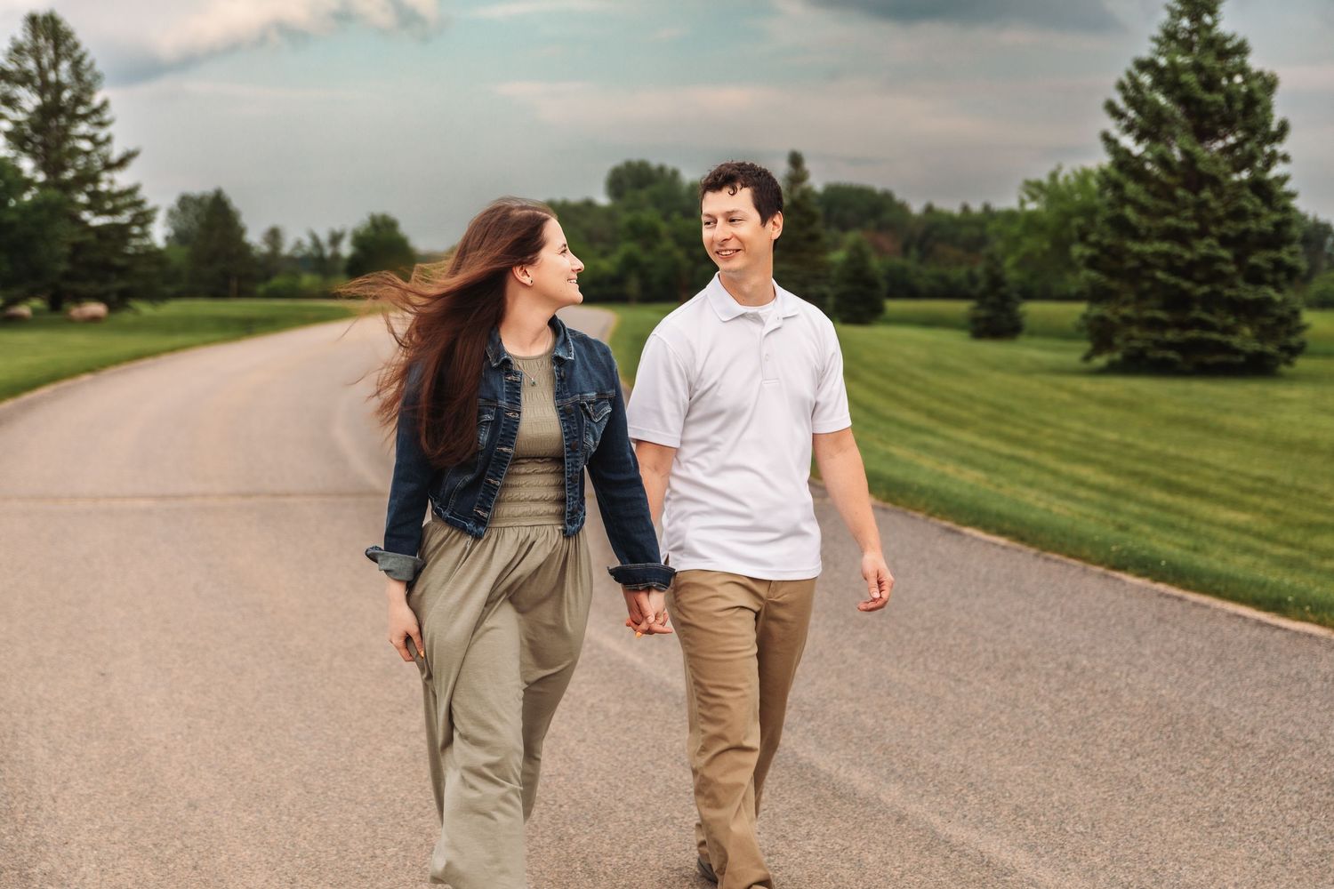 A couple walking and laughing together on a scenic country road with green lawns and evergreen trees in summer.