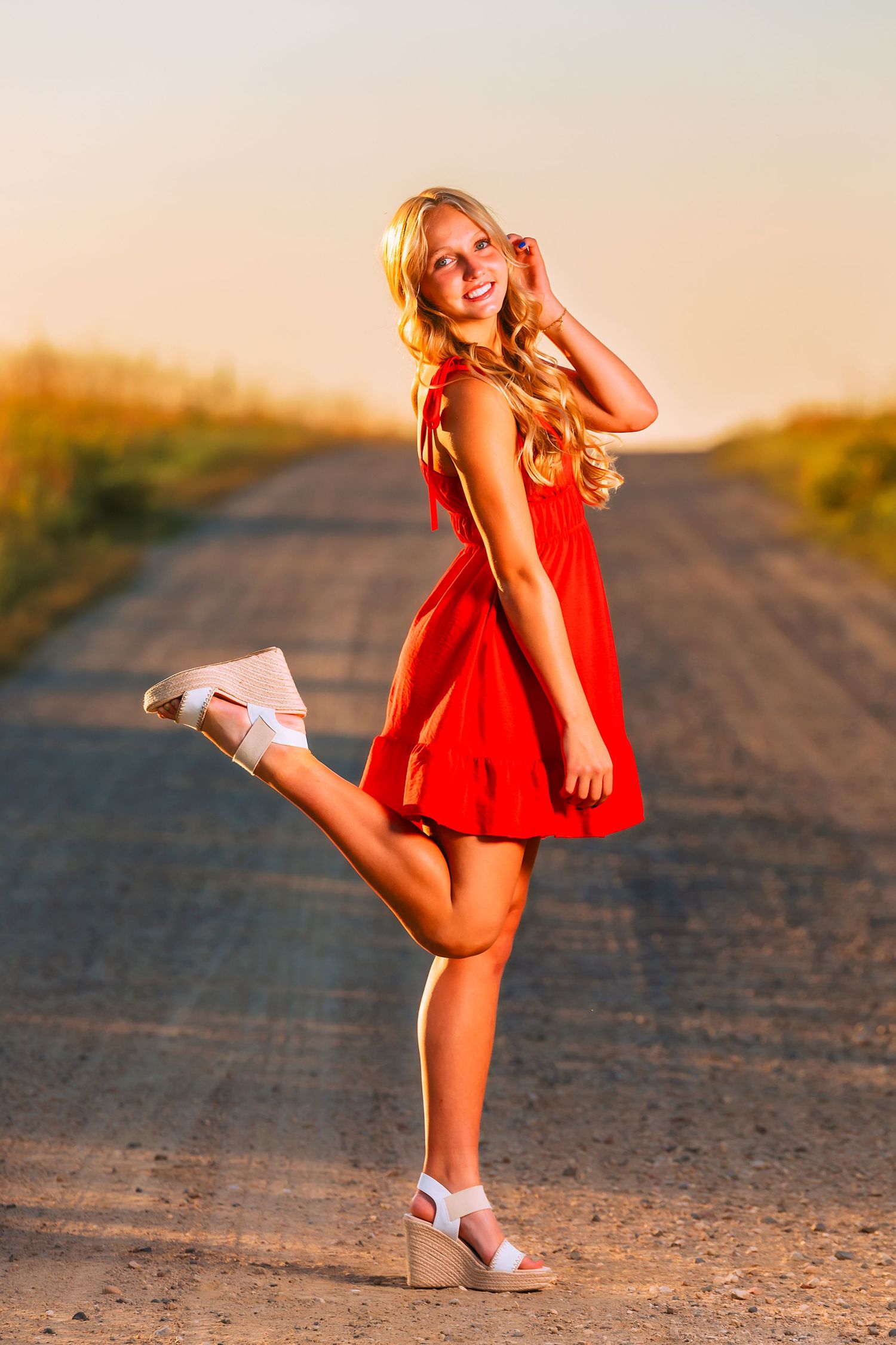 Portrait of someone in a red sundress striking a playful pose on a dirt road at sunset.