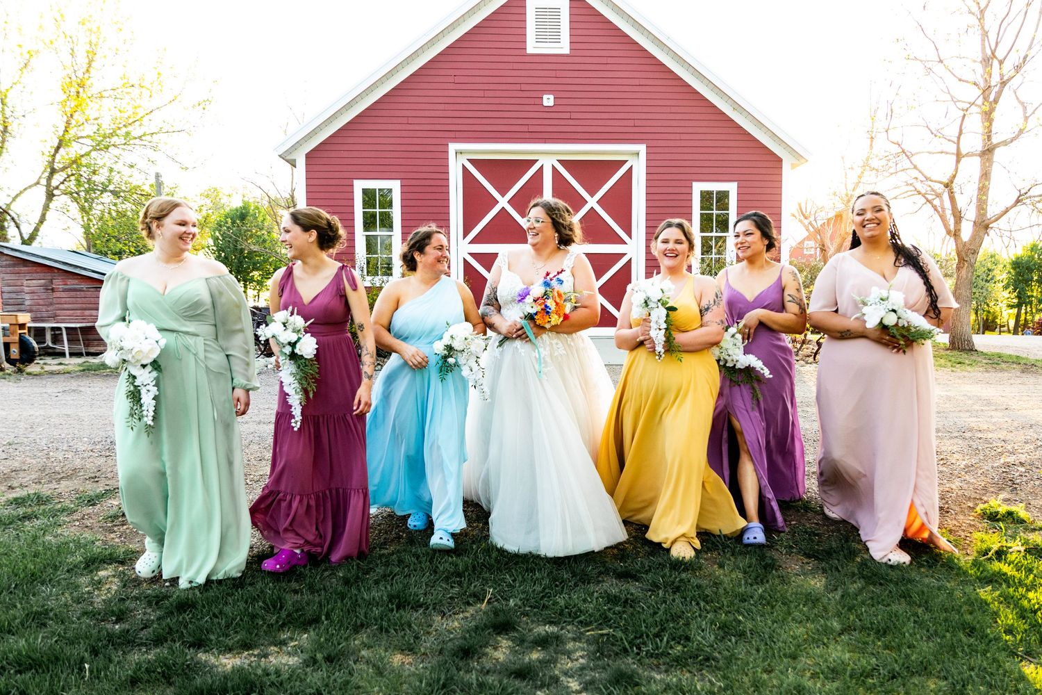 Rainbow colored bridesmaid dresses create a vibrant display against a classic red barn backdrop.