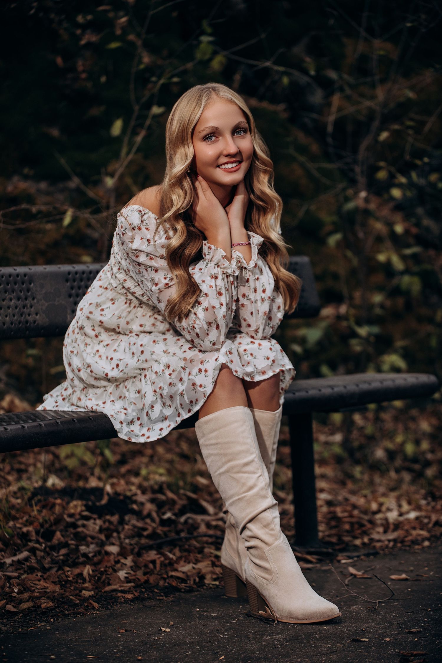 A person in a floral dress and cream boots sits on a bench surrounded by autumn foliage.