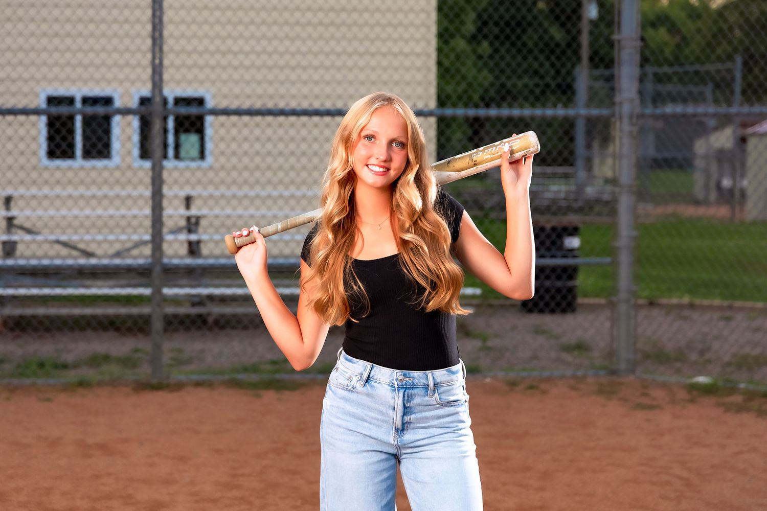 Athlete with a baseball bat poses on a dirt field at sunset wearing jeans and a black top.
