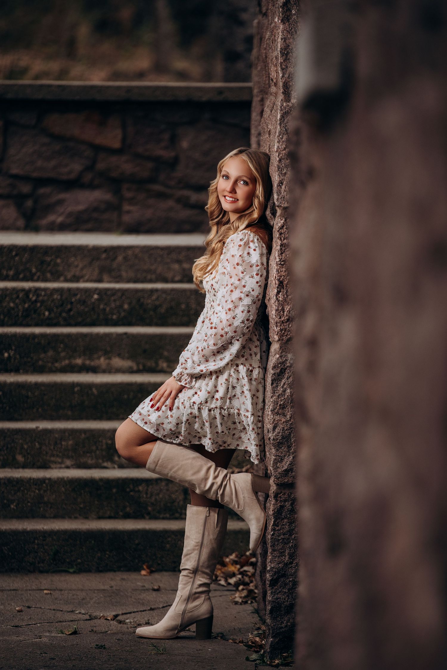 A model in a floral dress and tall boots poses on stone steps near an old wall.