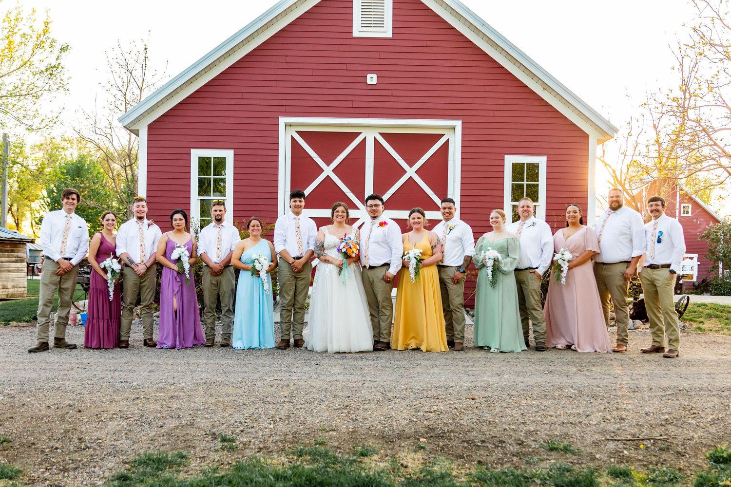 Wedding party poses in colorful attire in front of a classic red barn.