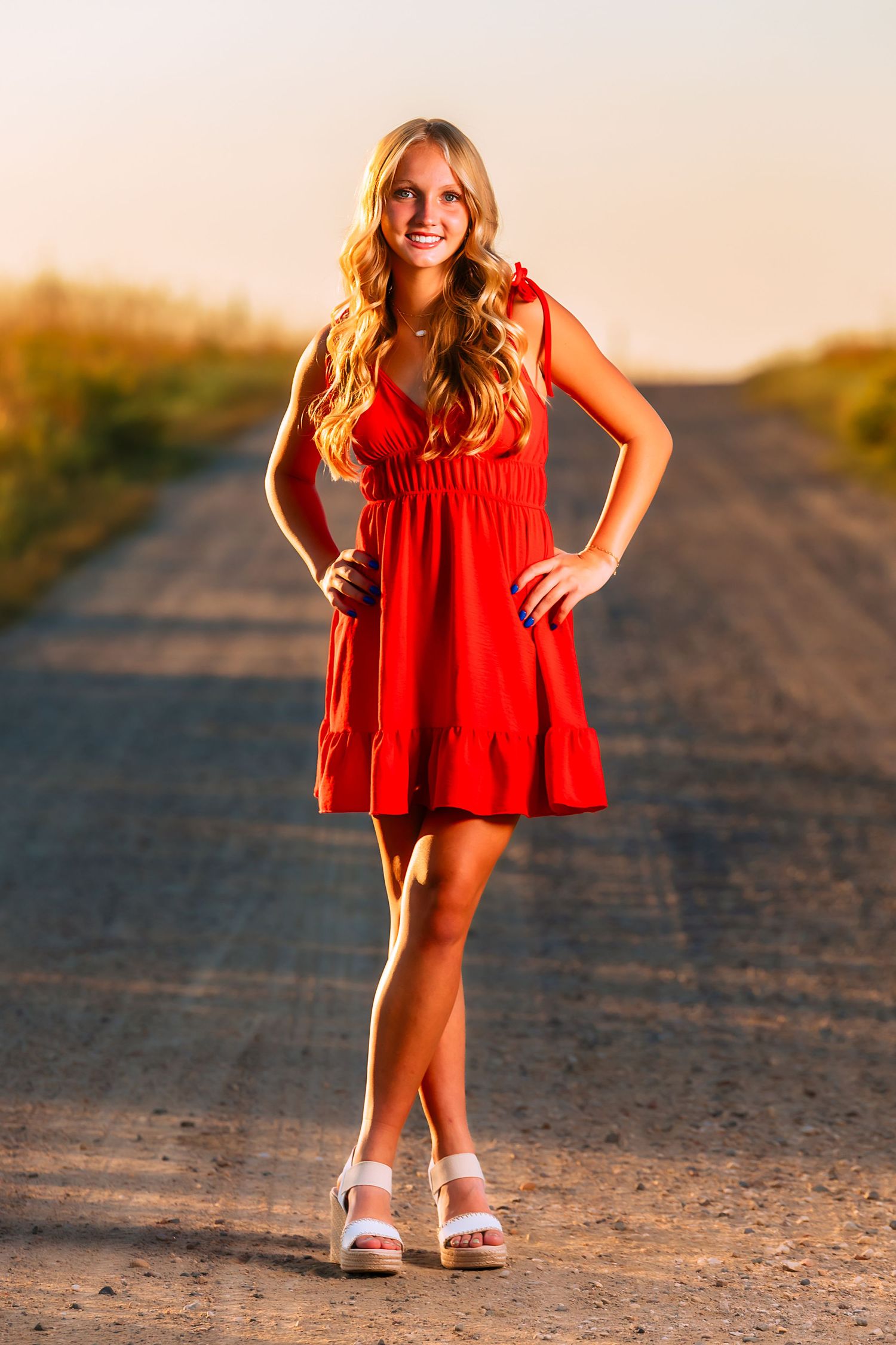 Senior portrait in red dress posed on dirt road at sunset.