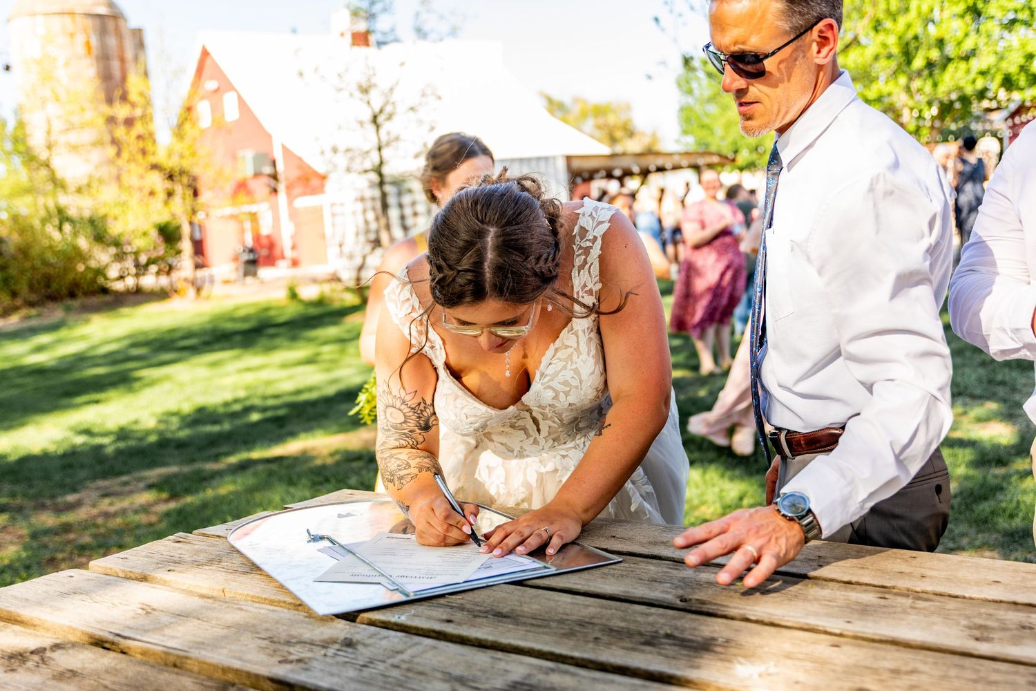 Wedding party members sign documents at an outdoor wooden table during a sunny day.
