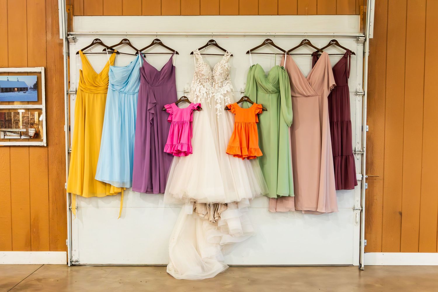 Wedding dress and colorful bridesmaid dresses hanging on display rack against orange wooden wall.