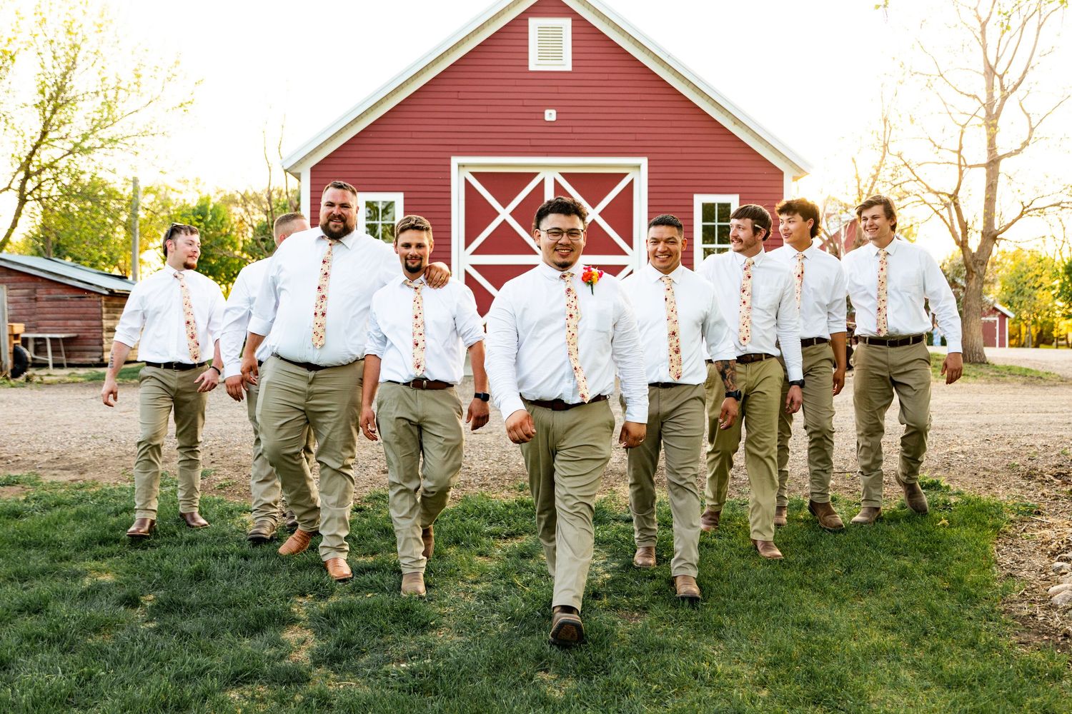 Groomsmen in khaki pants and white shirts walk in formation near a red barn.