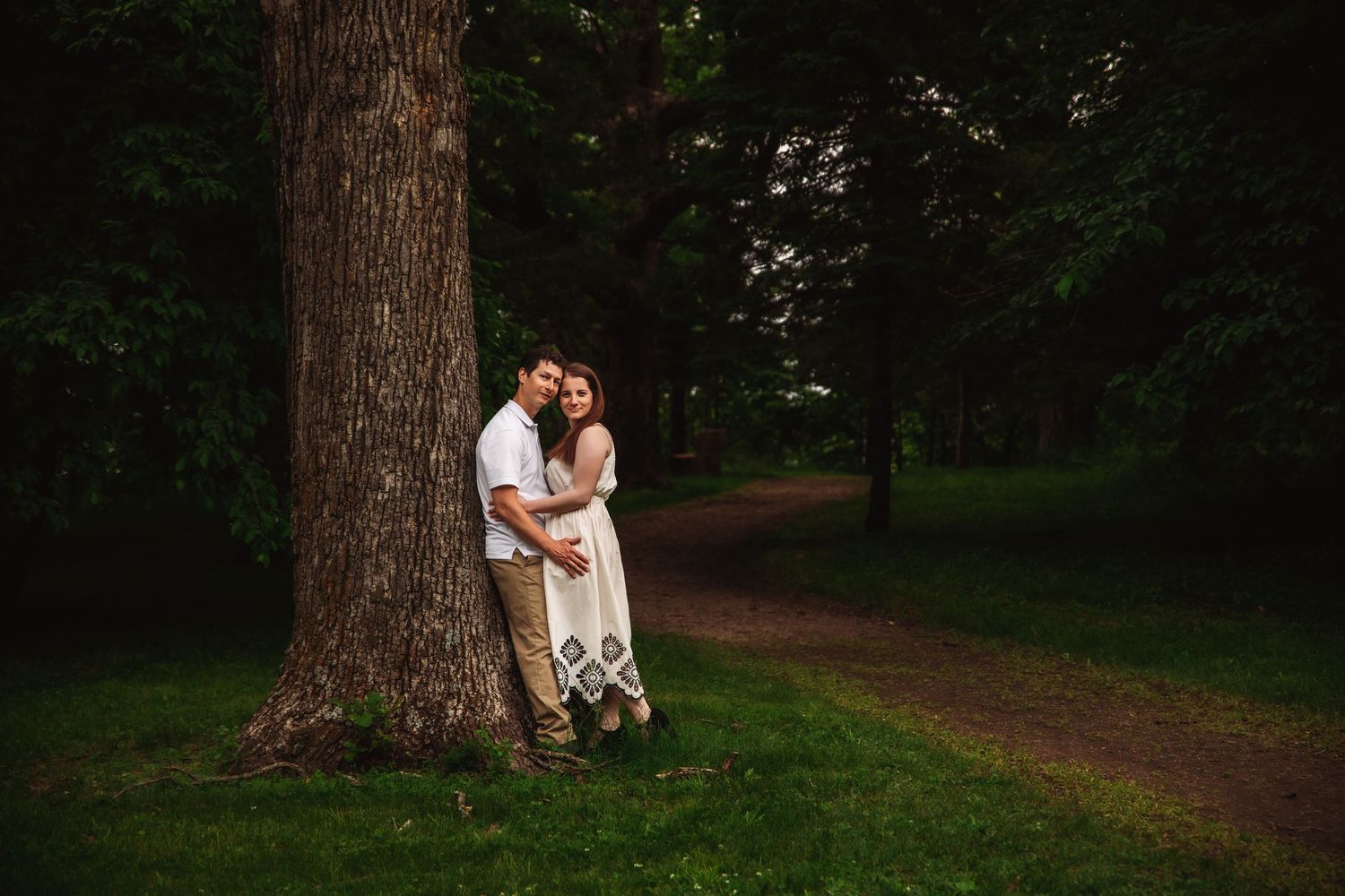 A couple shares an intimate moment against a large tree trunk on a forest path.