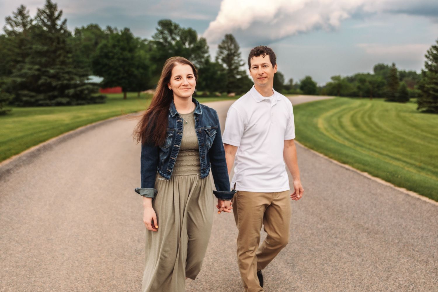 A couple walks hand in hand down a scenic country road with manicured grass and pine trees on a sunny day.