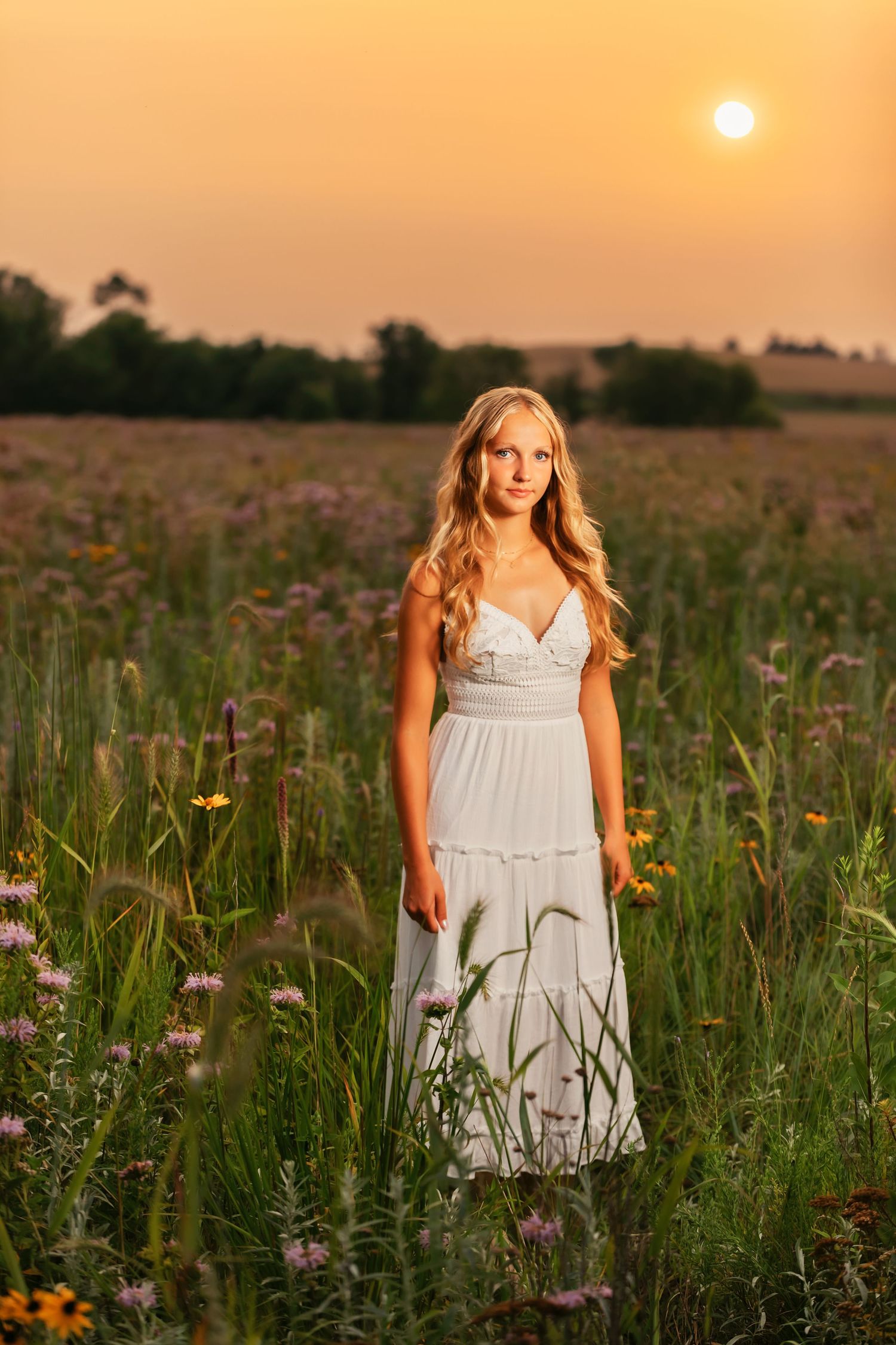 White dress catches sunset light while standing among blooming wildflowers in a meadow.